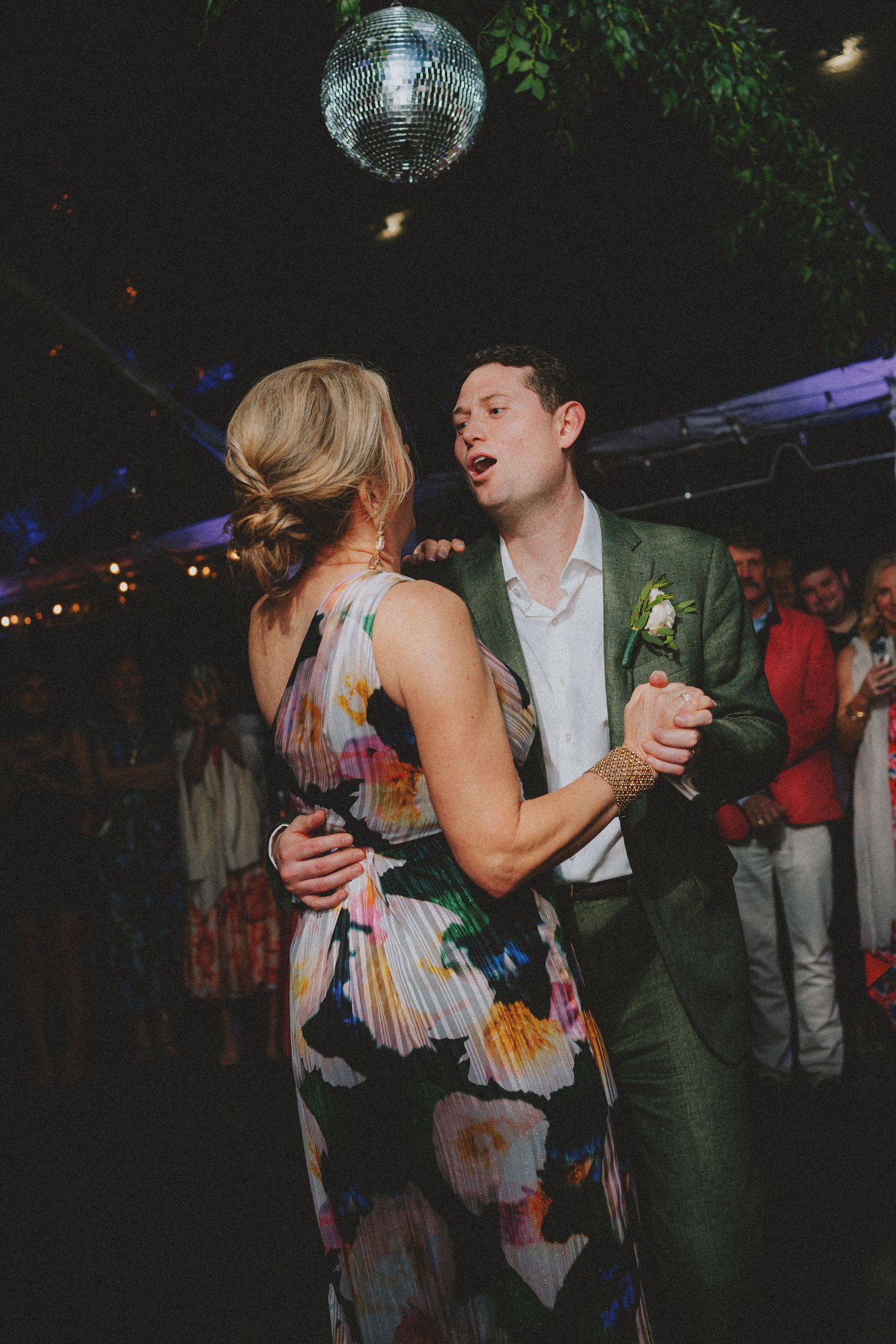 Groom in green suit dances with guest in floral dress under disco ball at wedding reception.