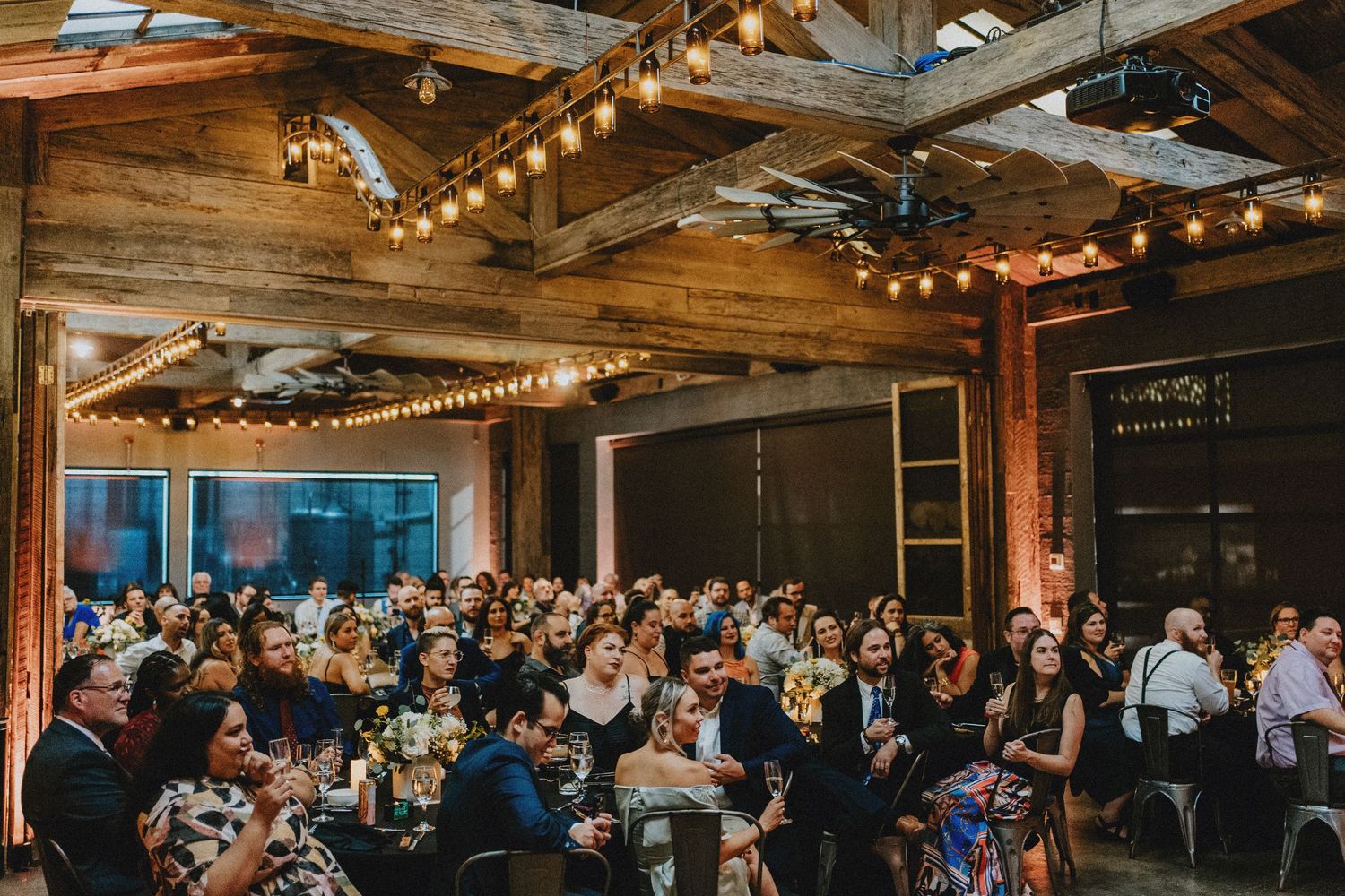 Rustic wedding reception venue with string lights, wooden beams, and guests seated at tables in a warm, industrial space.