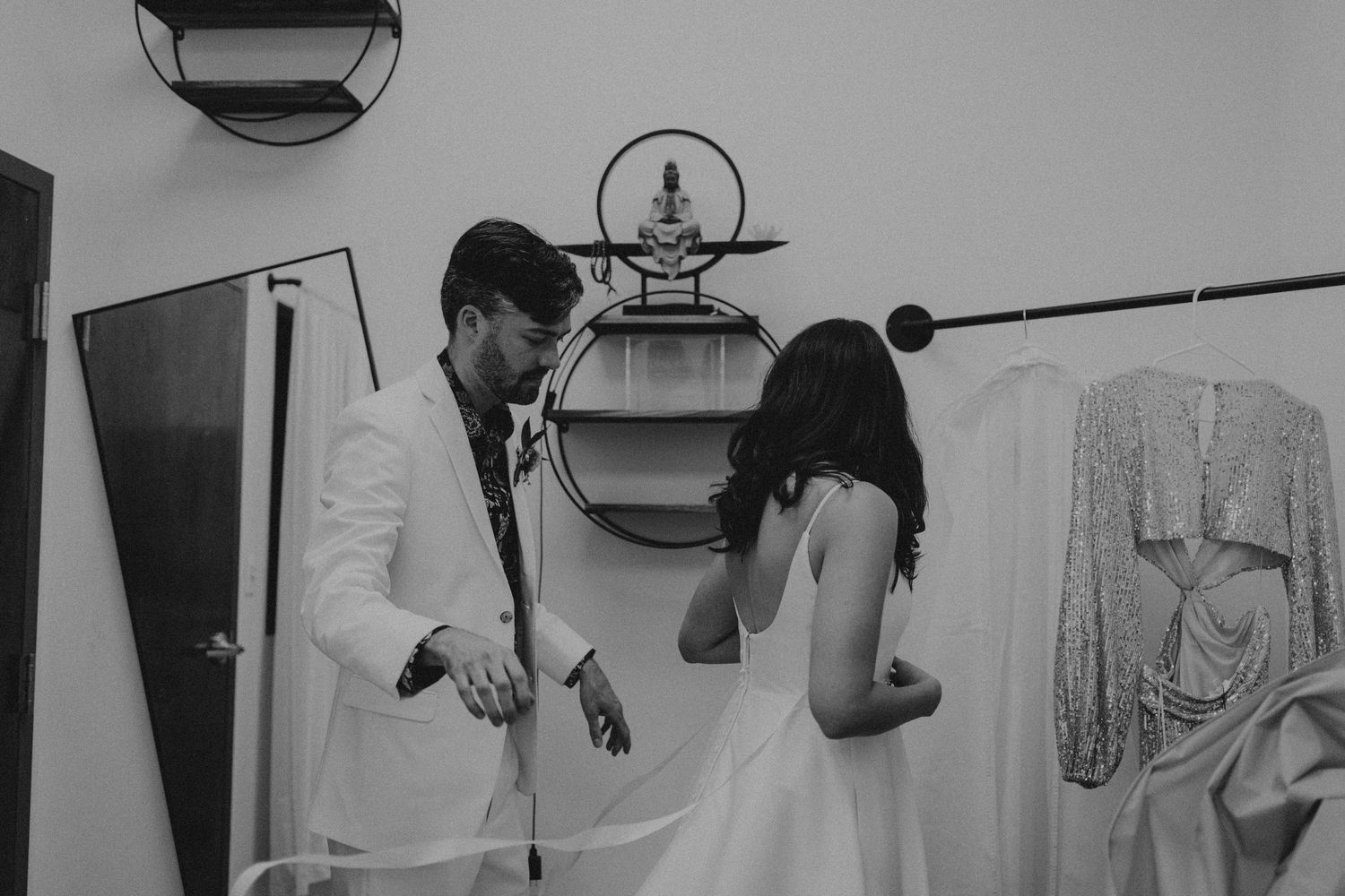 Groom in white suit helping bride with wedding dress in a modern black and white dressing room.