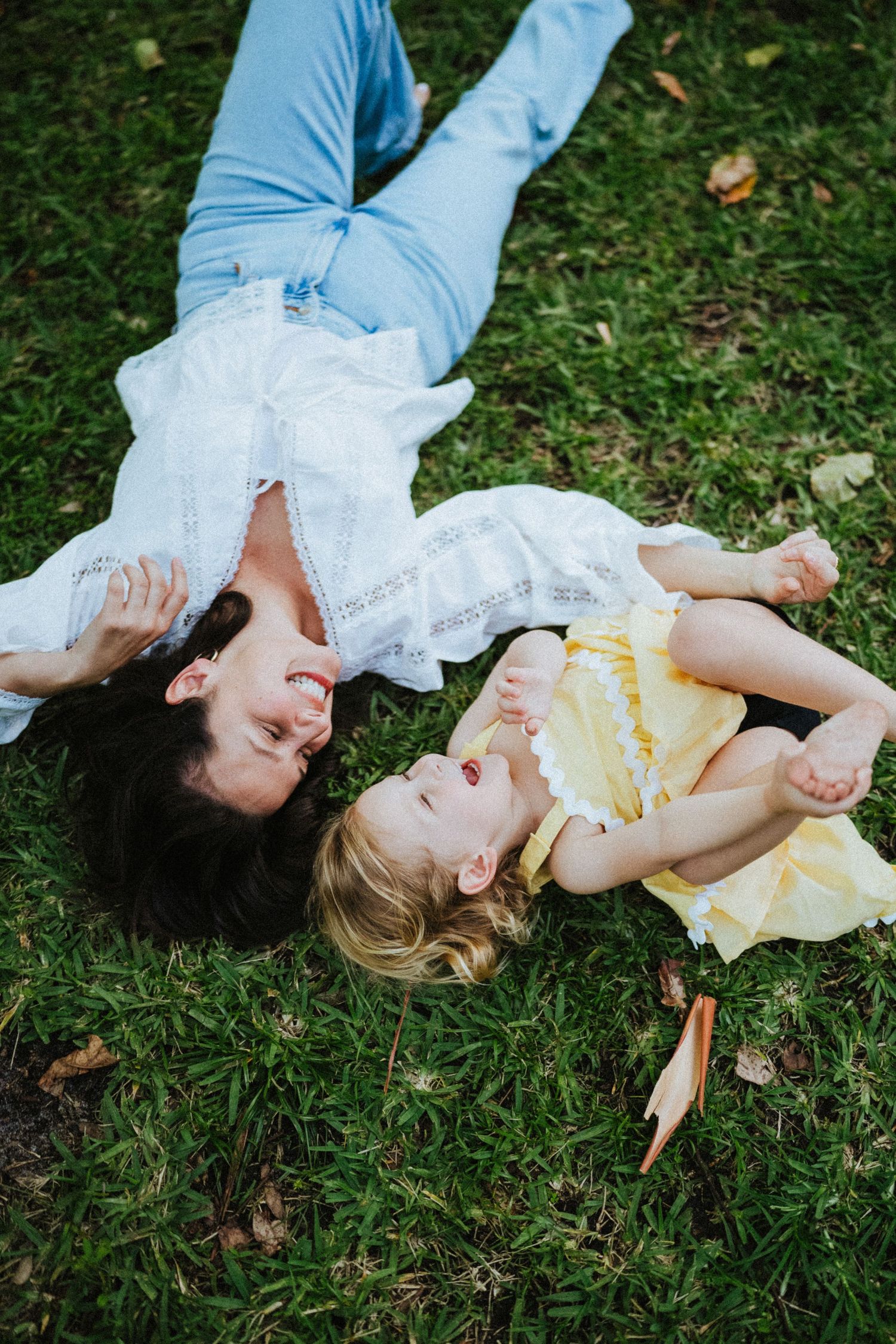 Mother and young daughter laughing together while lying on green grass, wearing white and yellow dresses.