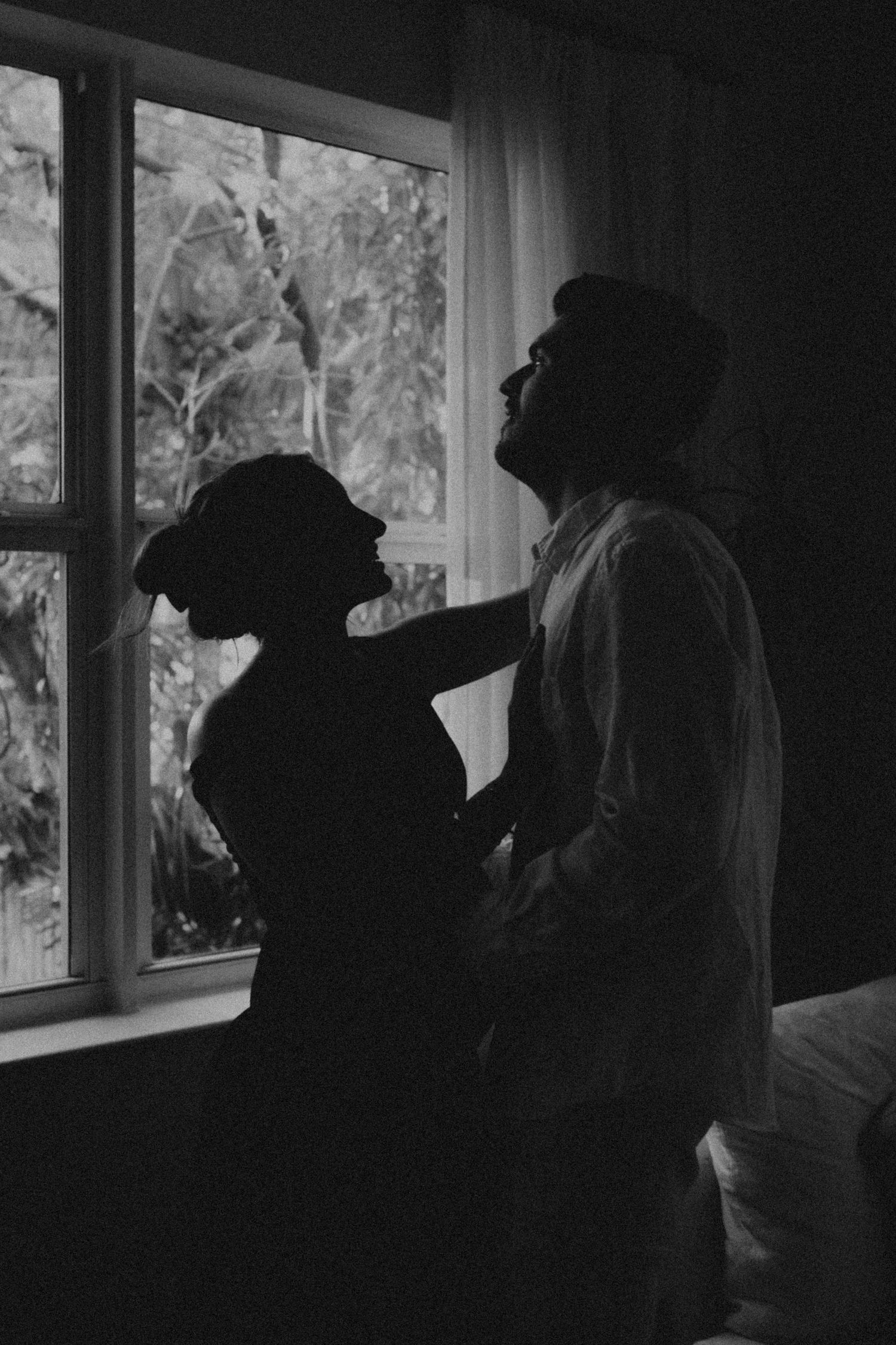 Black and white silhouette of a romantic couple embracing by a window with sheer curtains and trees outside.
