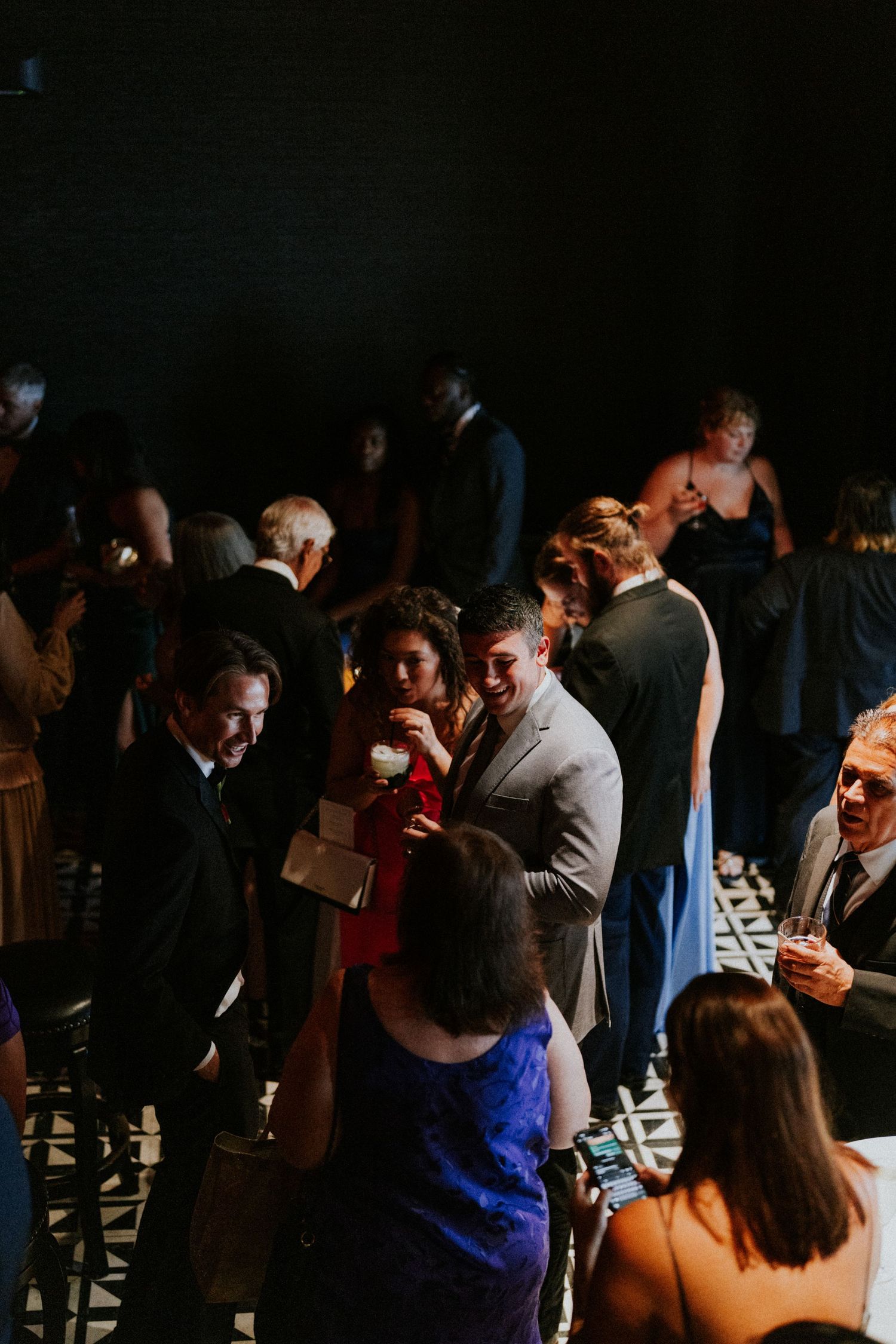 Wedding cocktail hour guests mingling and laughing in a dimly lit modern venue space.