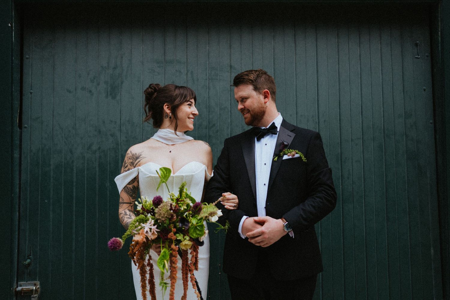 Bride and groom smiling at each other holding floral bouquet against green barn door on wedding day.