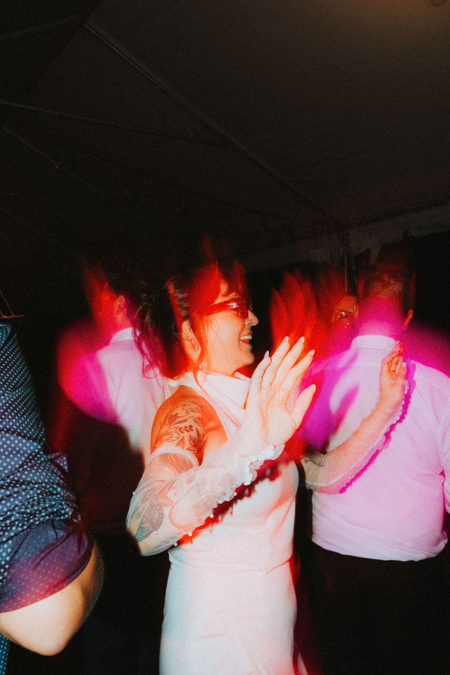 A bride in a white dress dances joyfully with guests under pink lights at a wedding reception party.