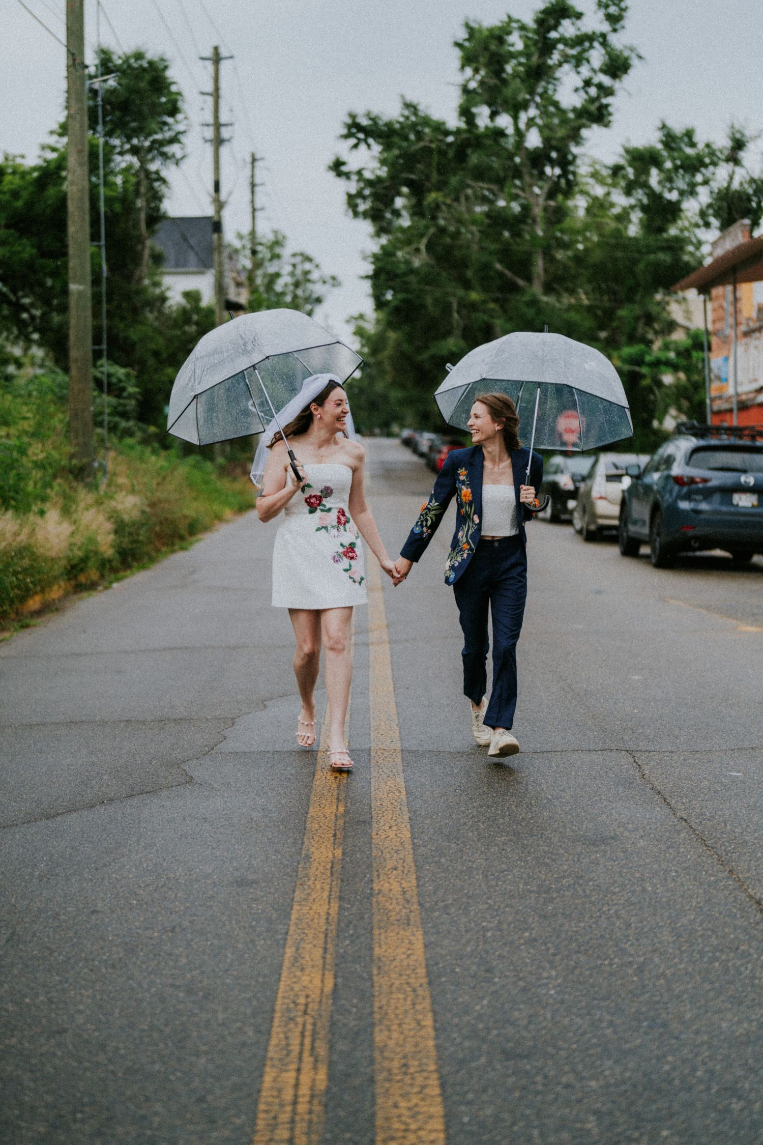 Two women holding hands and clear umbrellas walk together down a rain-slicked road lined with trees and parked cars.