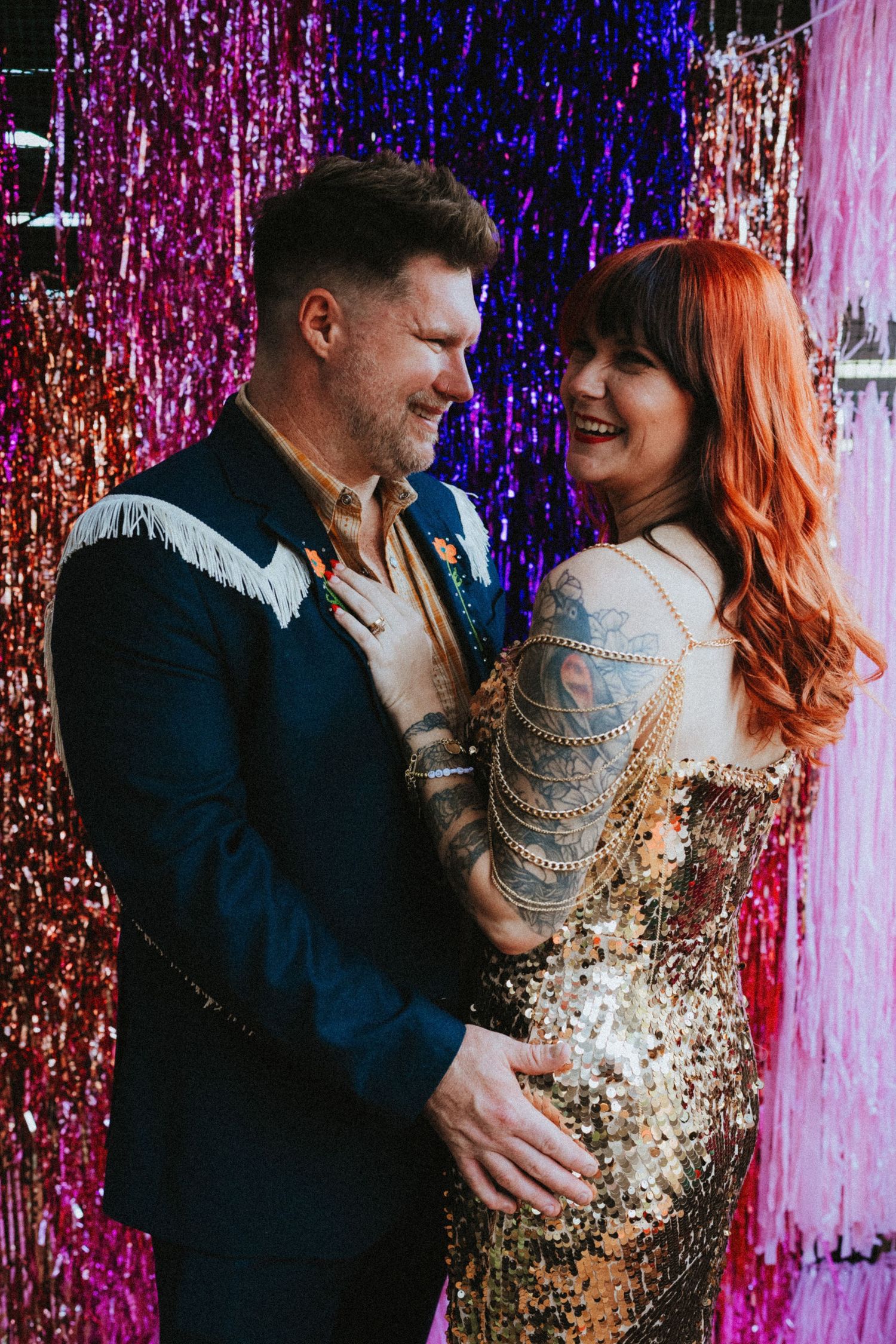 Couple in sequin and fringe outfits smiling together in front of colorful tinsel curtain backdrop.