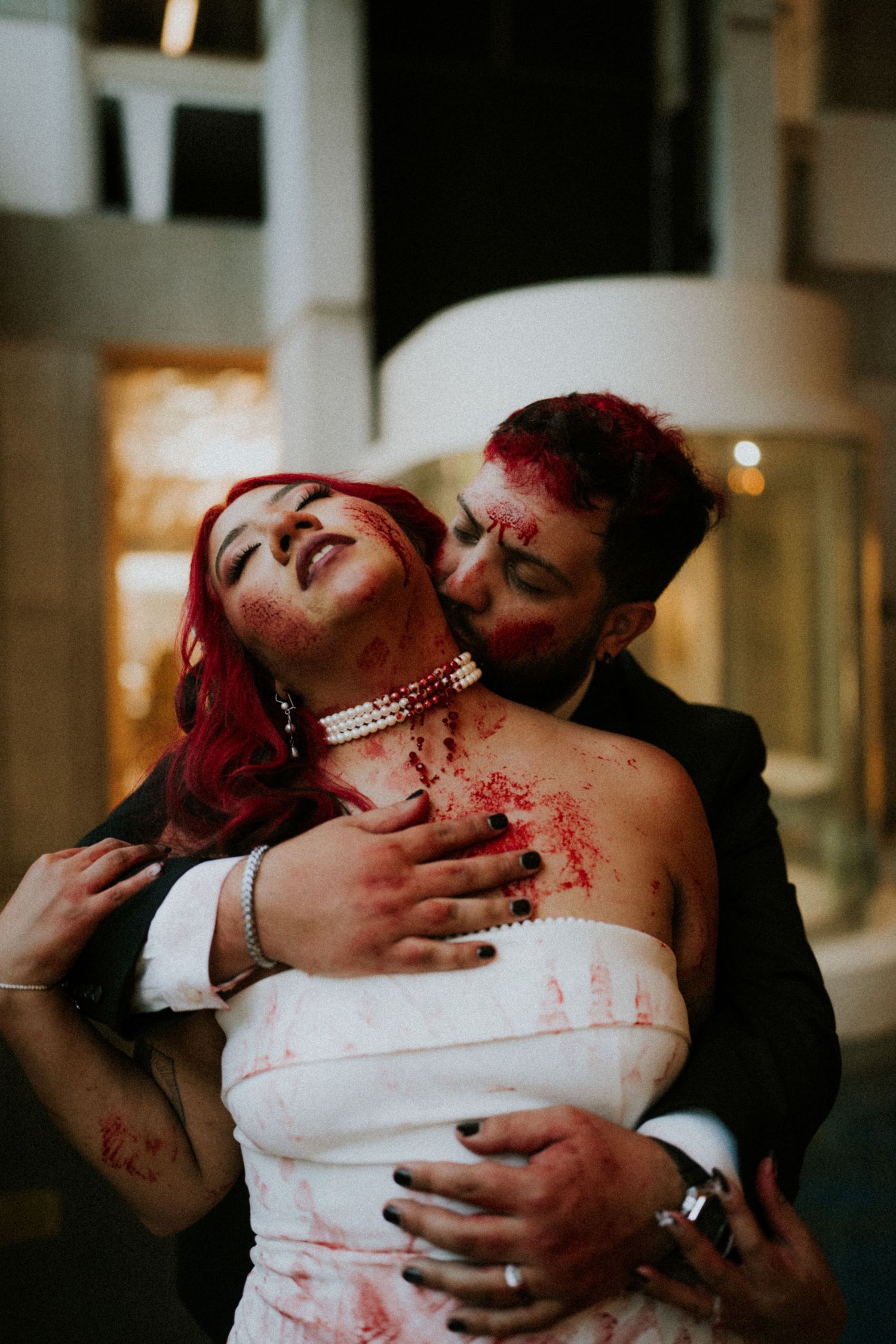 Couple in zombie wedding costumes with fake blood, bride in white dress with red hair being embraced by groom in suit.