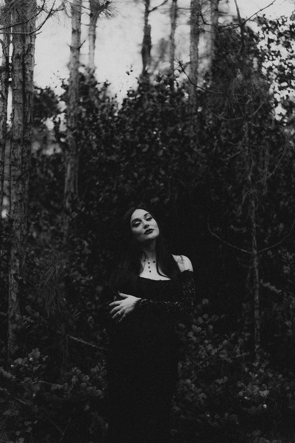 Dramatic black and white portrait of woman in dark dress among forest trees.