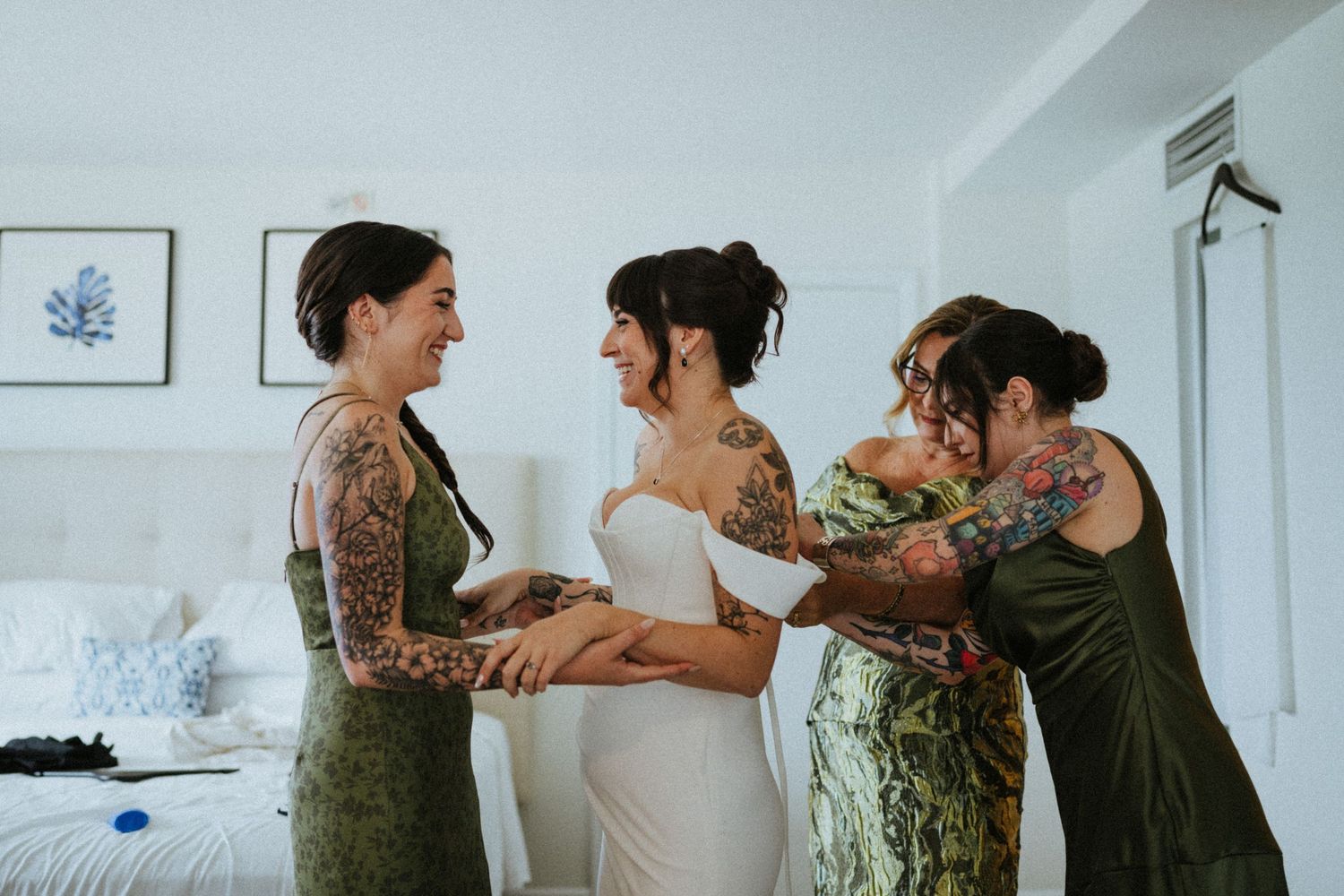 Bride in white gown being helped by tattooed bridesmaids in green dresses in a bright white bedroom.