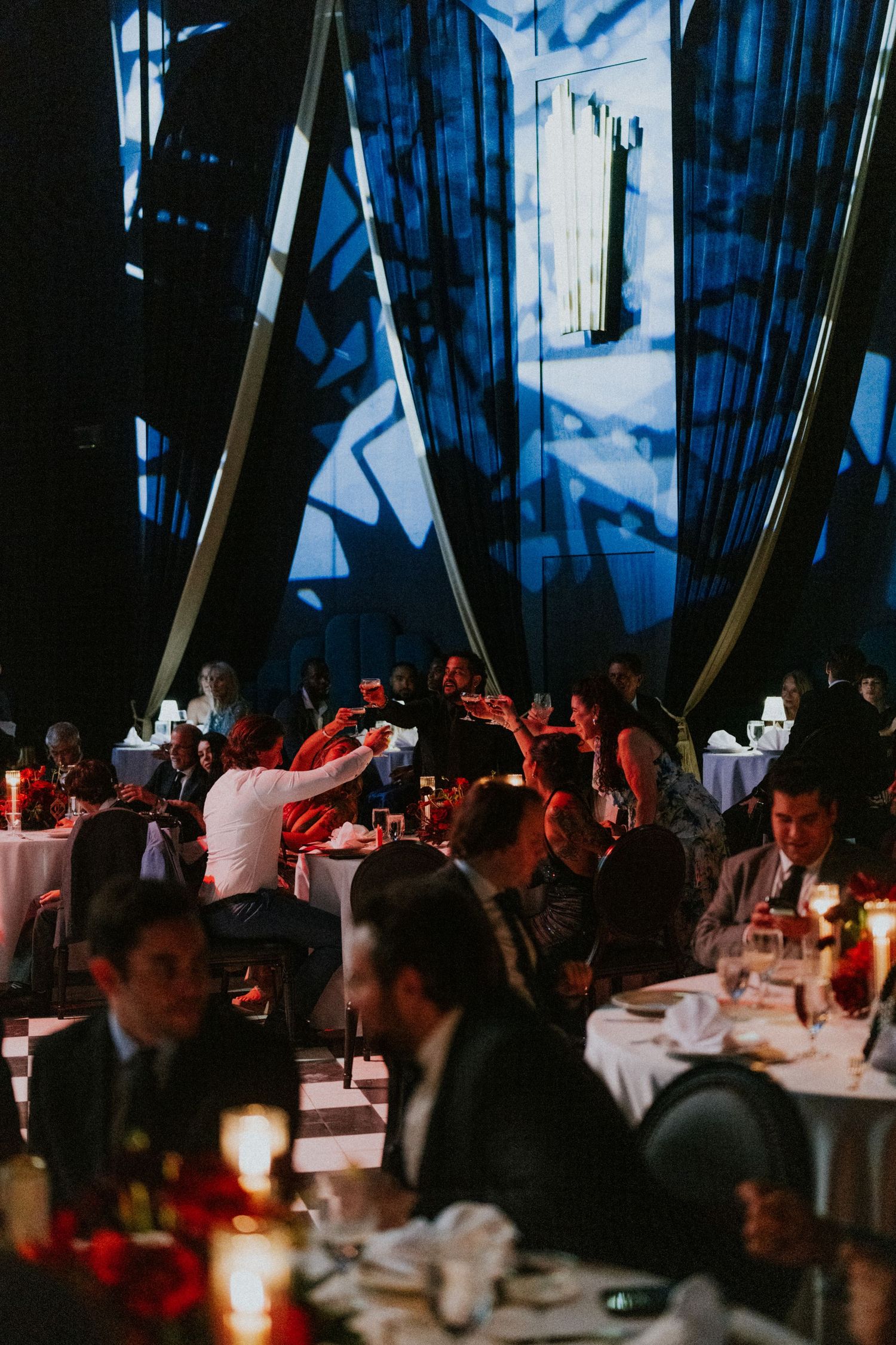 Elegant candlelit wedding reception dinner in a dramatic venue with blue projected lighting and guests raising glasses.
