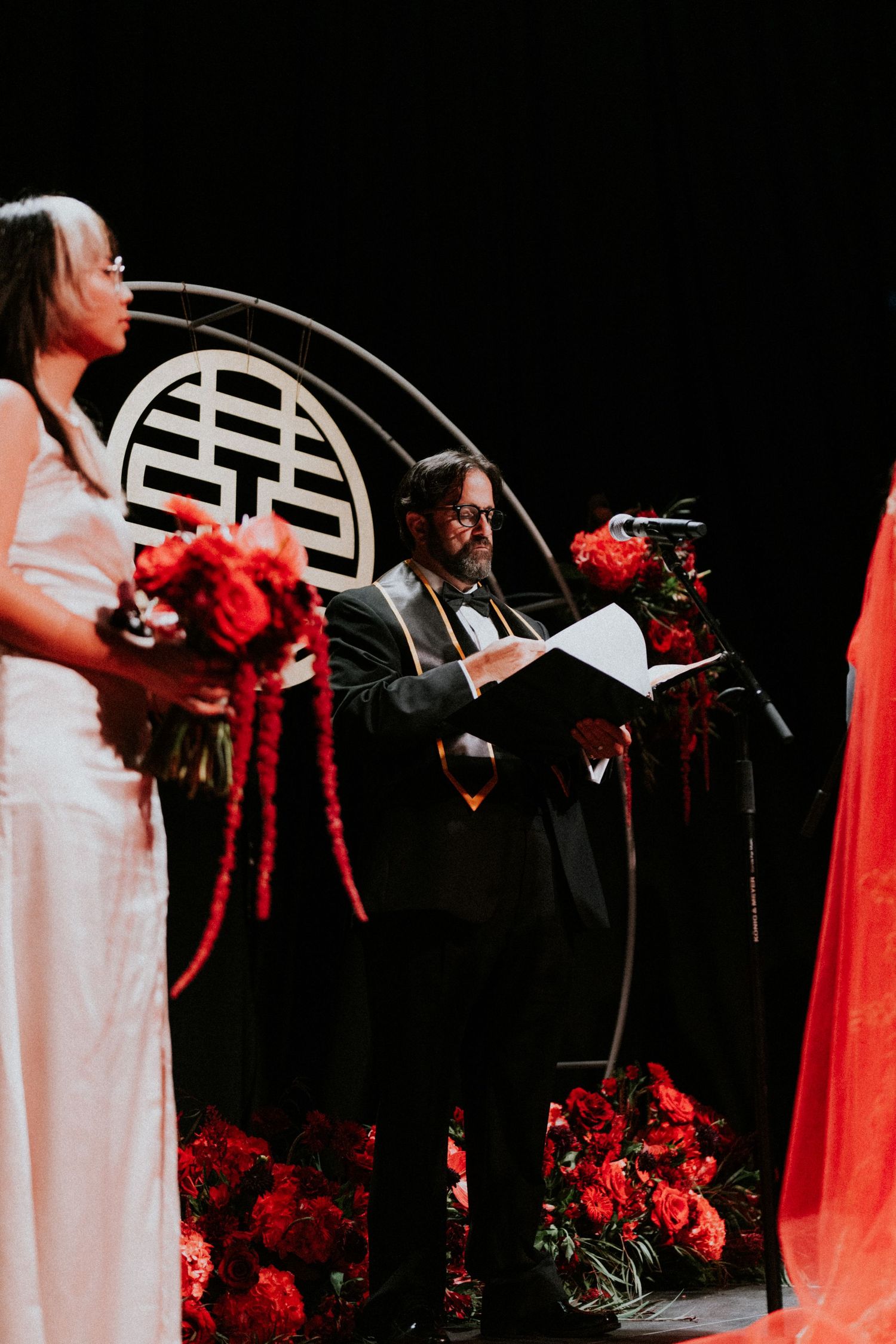 Officiant reads vows at elegant wedding ceremony with red floral arrangements and Chinese double happiness symbol backdrop.
