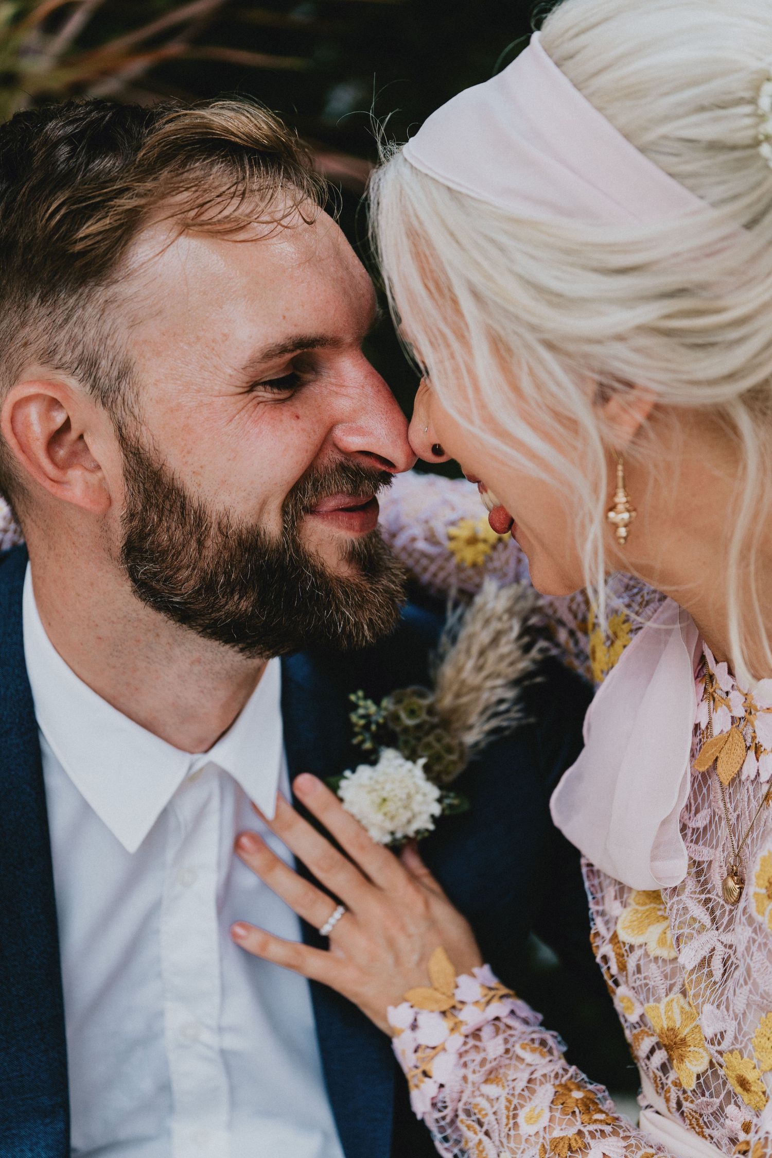 A bearded groom in a blue suit and blonde bride in floral lace dress share a romantic nose-to-nose moment on their wedding day.