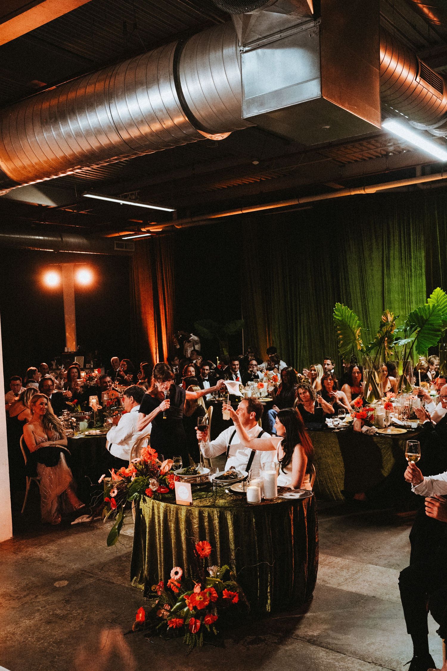 Wedding reception toast in an industrial venue with green drapes, tropical florals, and warm amber lighting.