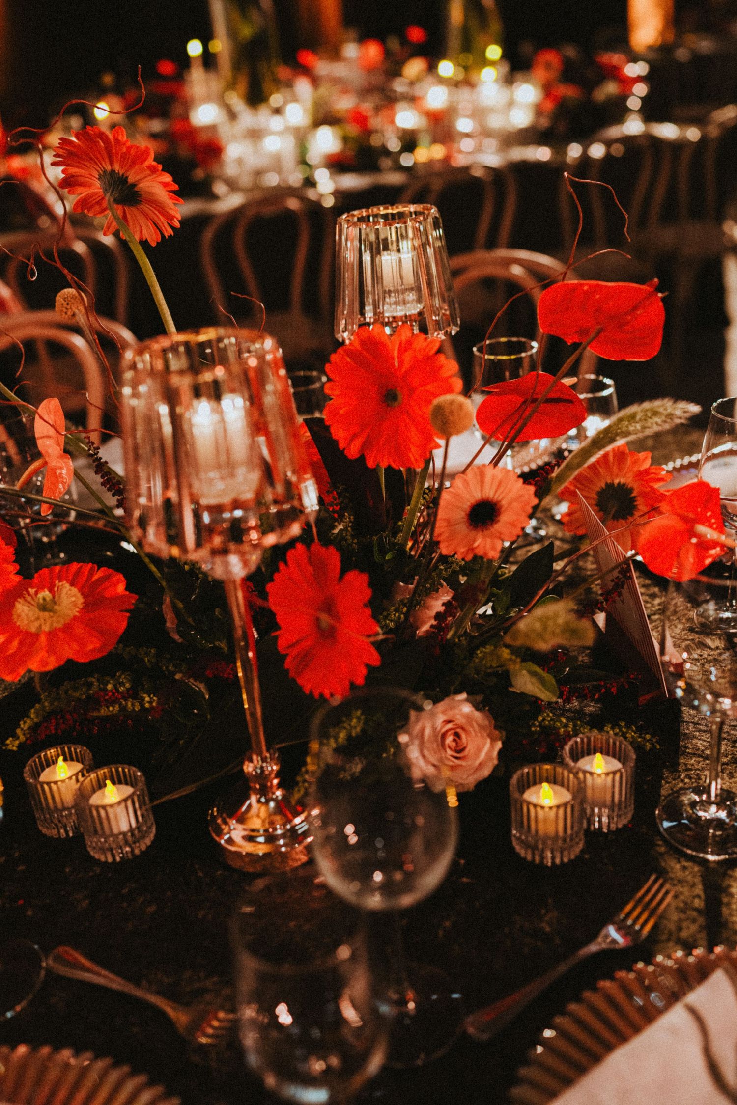 Elegant wedding table centerpiece with vibrant red gerbera daisies, crystal candle holders, and glowing tea lights on a dark surface.