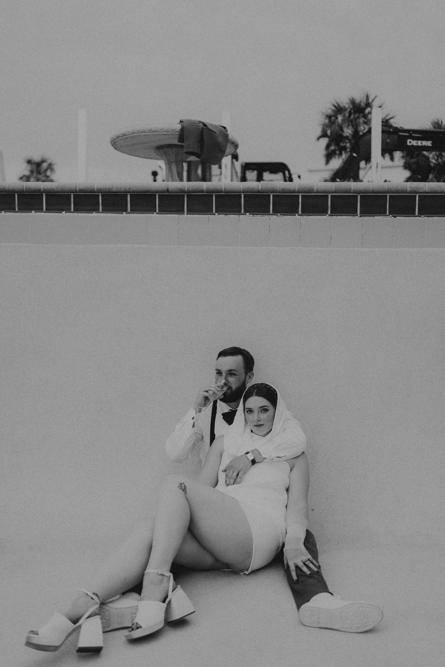 Couple in wedding attire sitting together on the ground in a black and white outdoor photo.