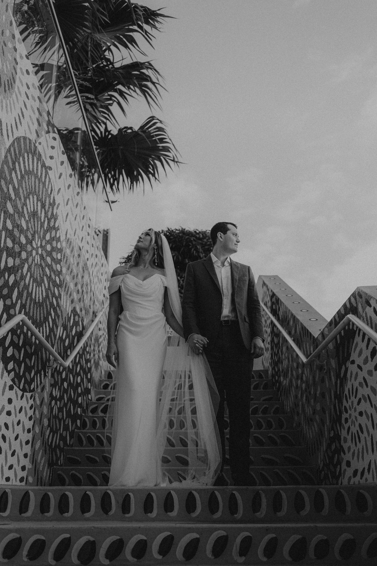 Bride and groom holding hands on decorated staircase with palm tree in black and white.