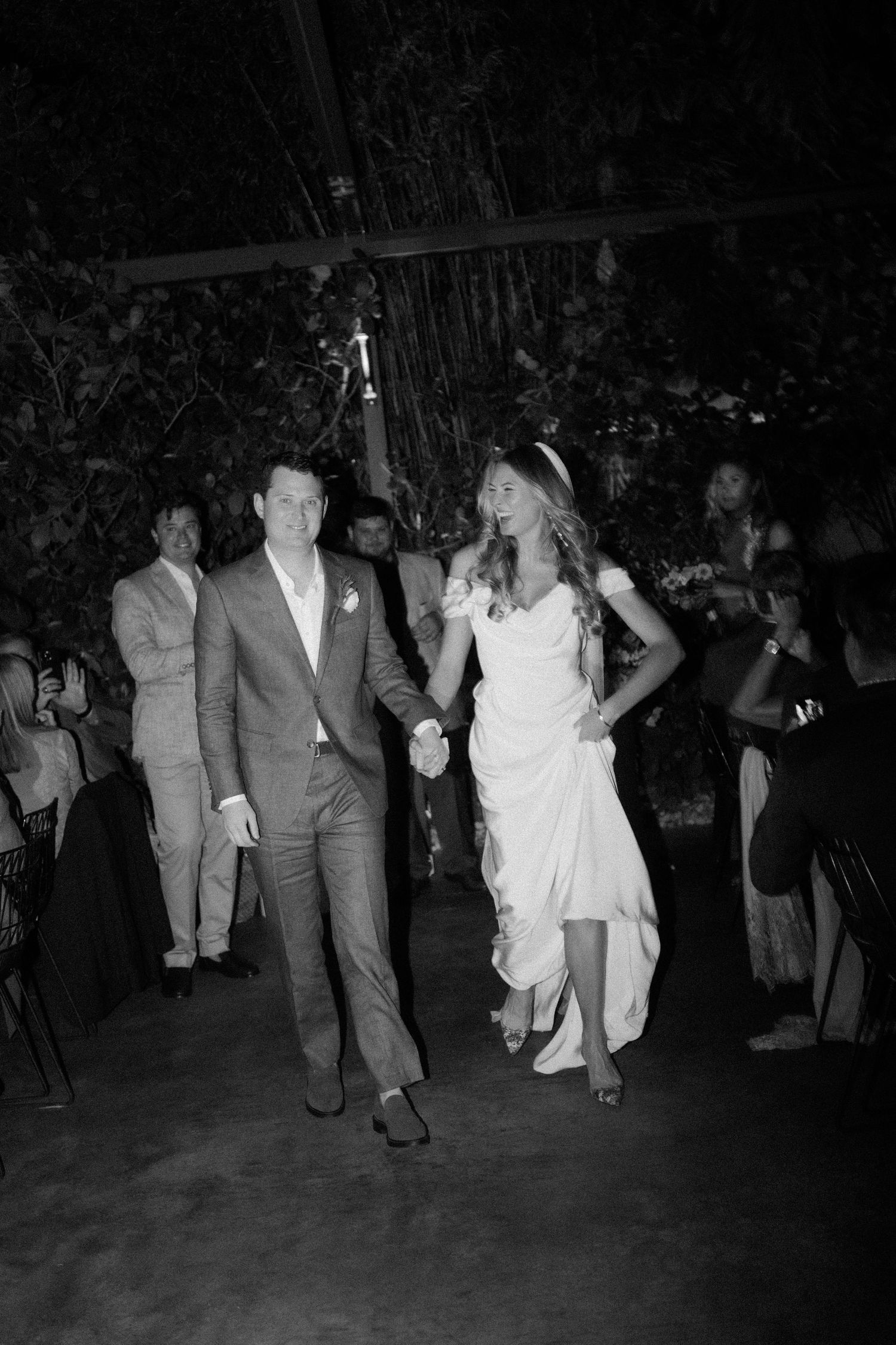 Black and white photo of a bride and groom holding hands during their wedding reception entrance.