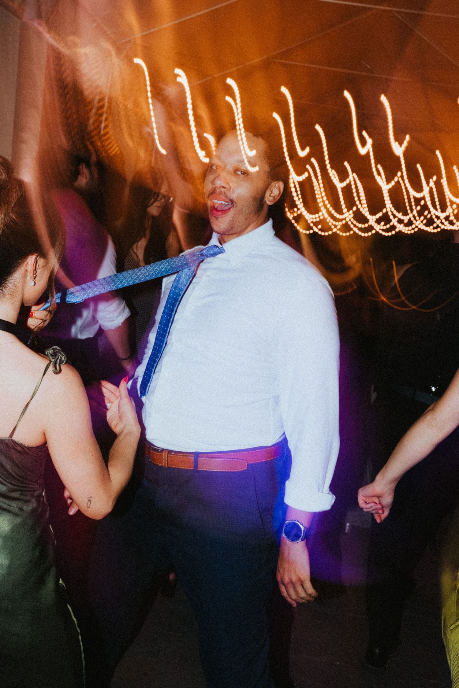 Energetic wedding guest dances with tie flying at a lively reception under warm string lights.