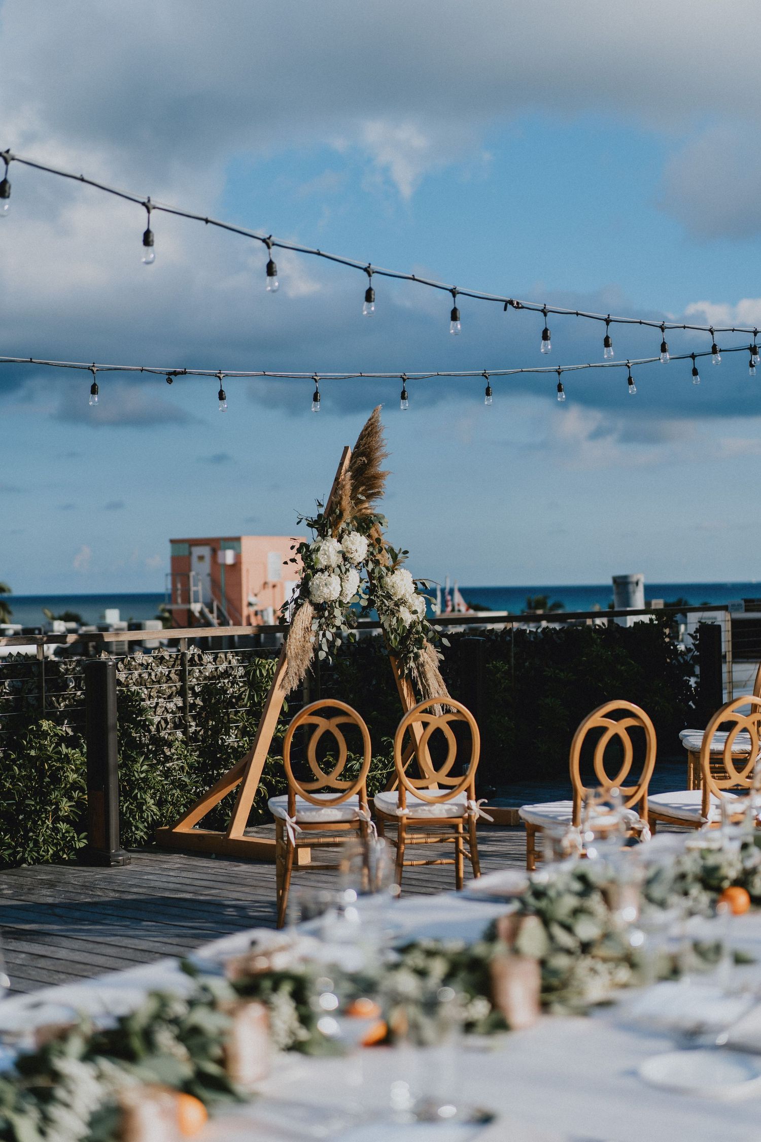 Outdoor rooftop wedding reception with rattan chairs, floral arch, string lights, and ocean view under cloudy sky.