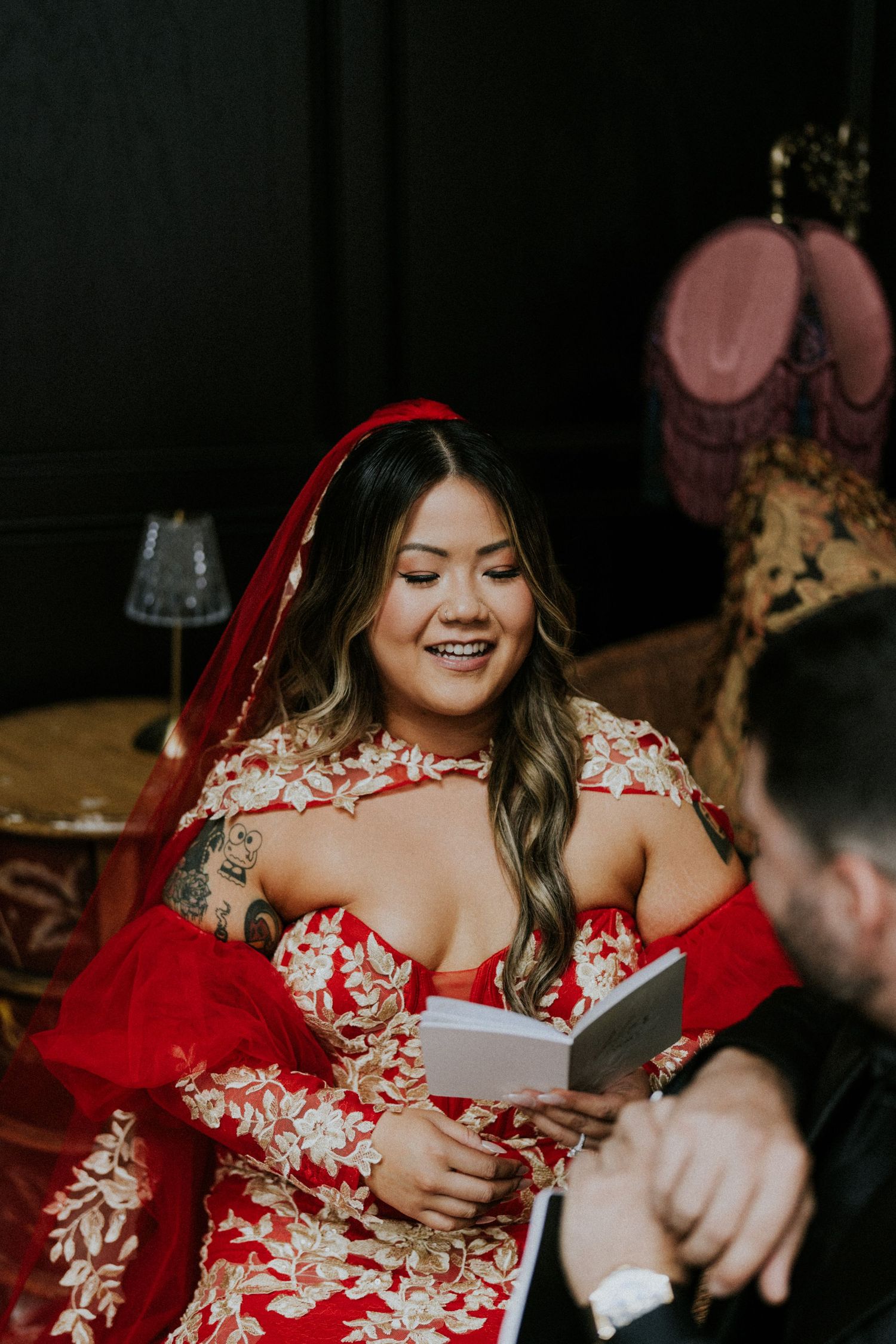 Smiling bride in red and gold embroidered wedding gown with red veil reads vows at indoor ceremony.