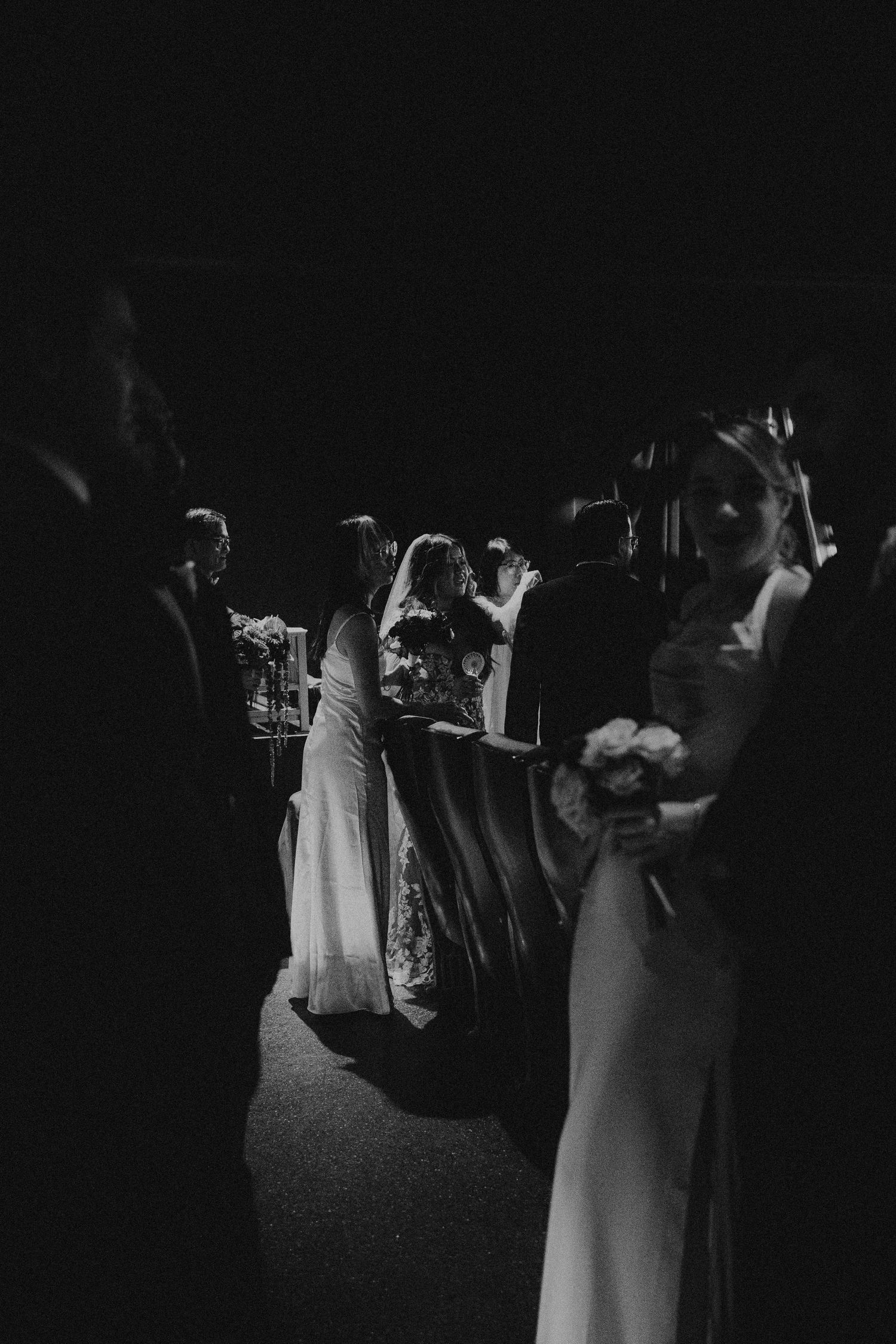 Black and white photo of elegantly dressed wedding guests in white gowns socializing at a dimly lit reception.