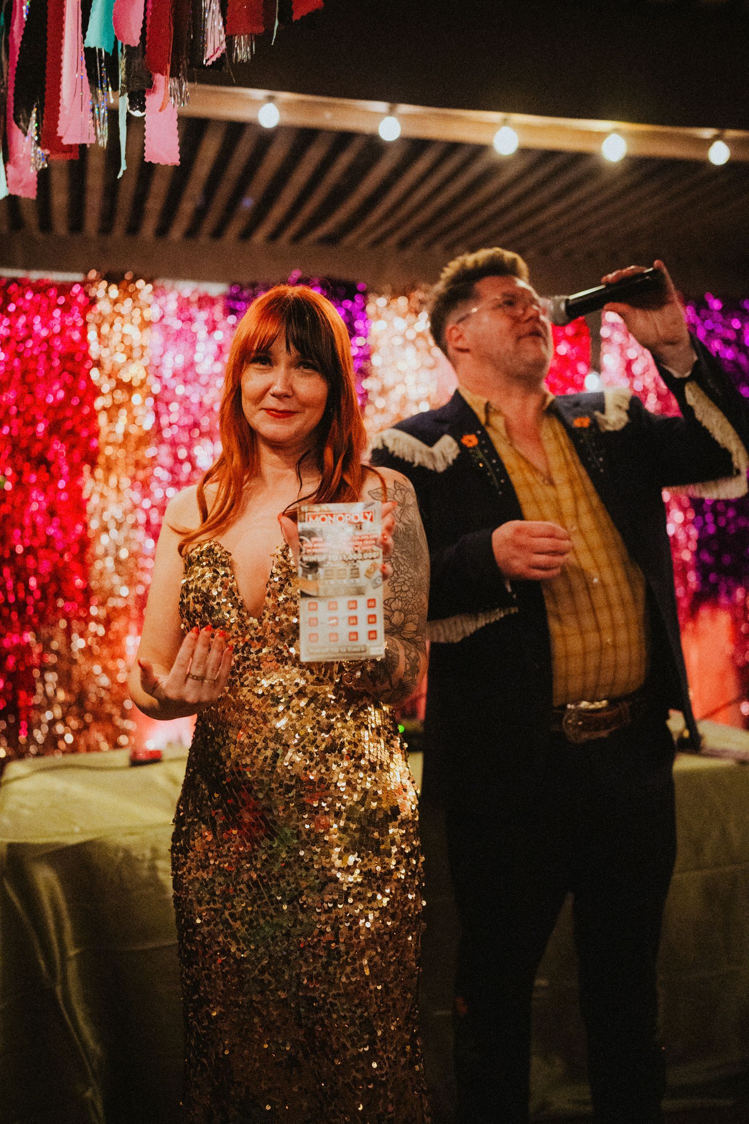 Woman in gold sequin dress holds bingo card at colorful party with sequin backdrop and string lights.
