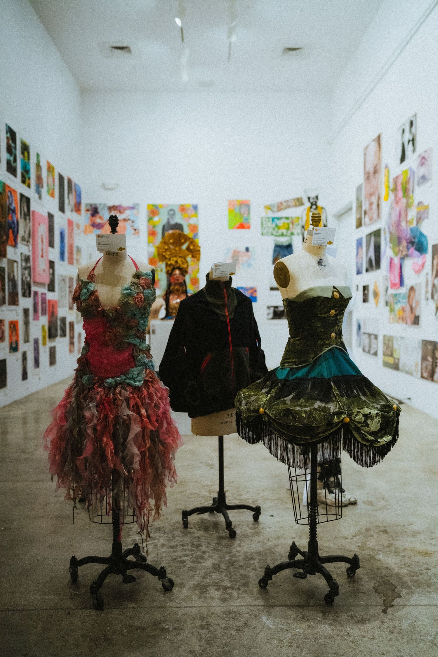 Three ornate dress forms displayed in an art gallery with colorful artwork covering white walls in the background.