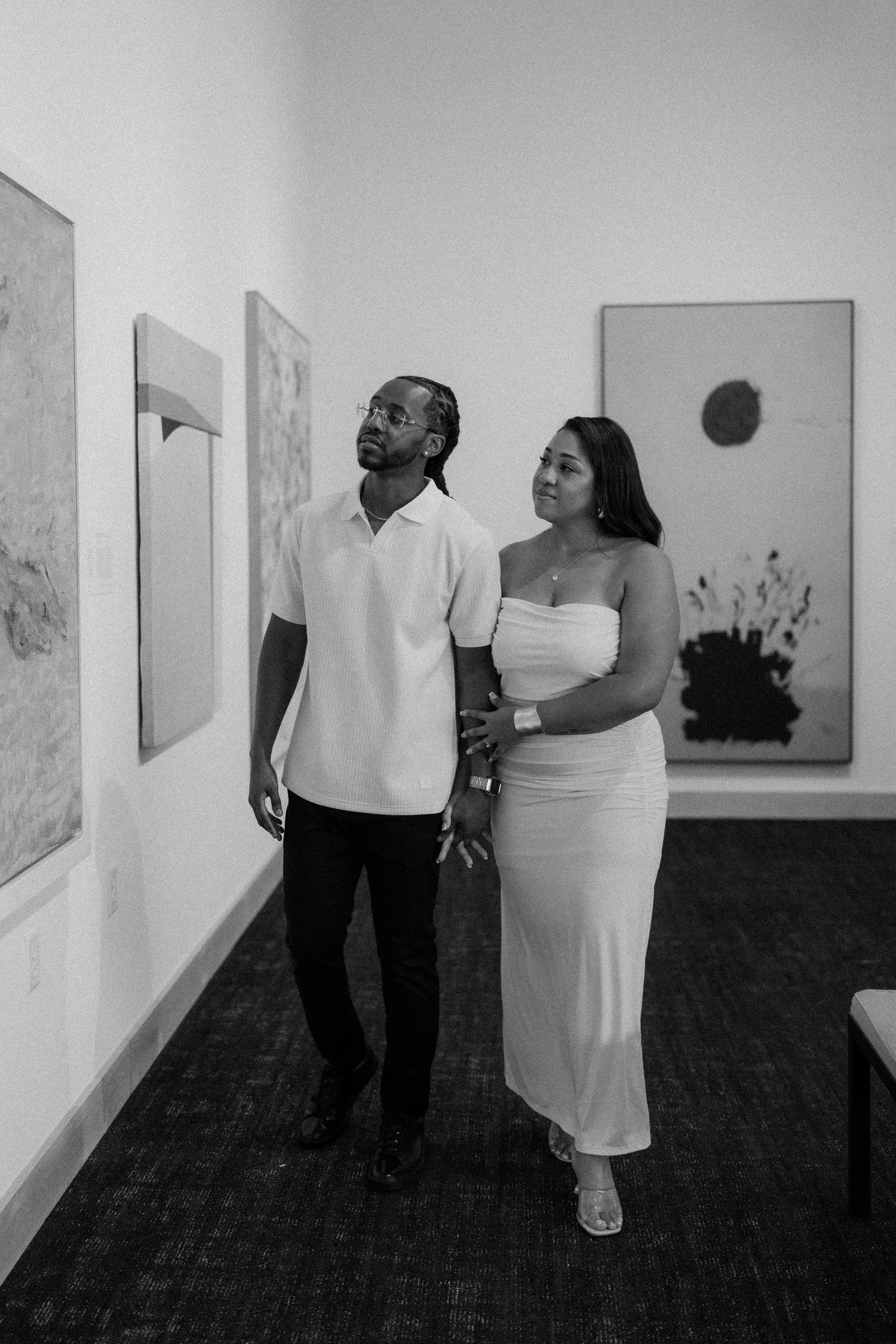A couple holds hands while admiring artwork in a modern gallery, dressed in white outfits, captured in black and white.