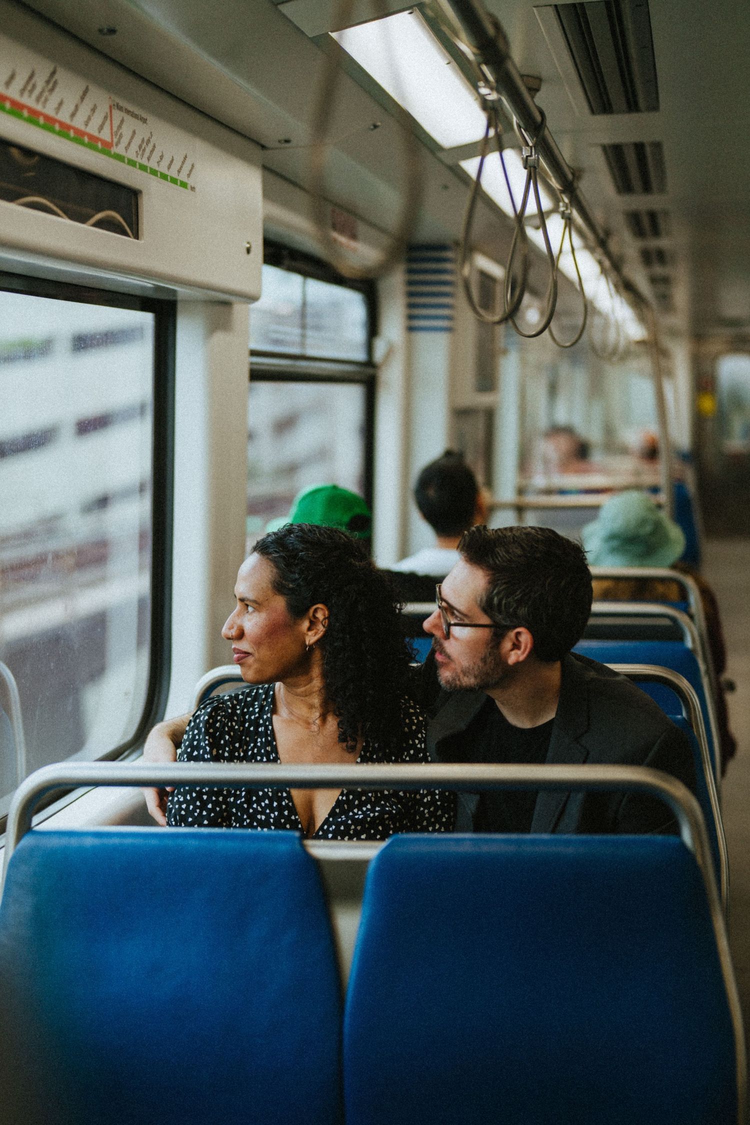 Couple sitting together on a train, woman gazing out window while man looks at her, blue seats visible.