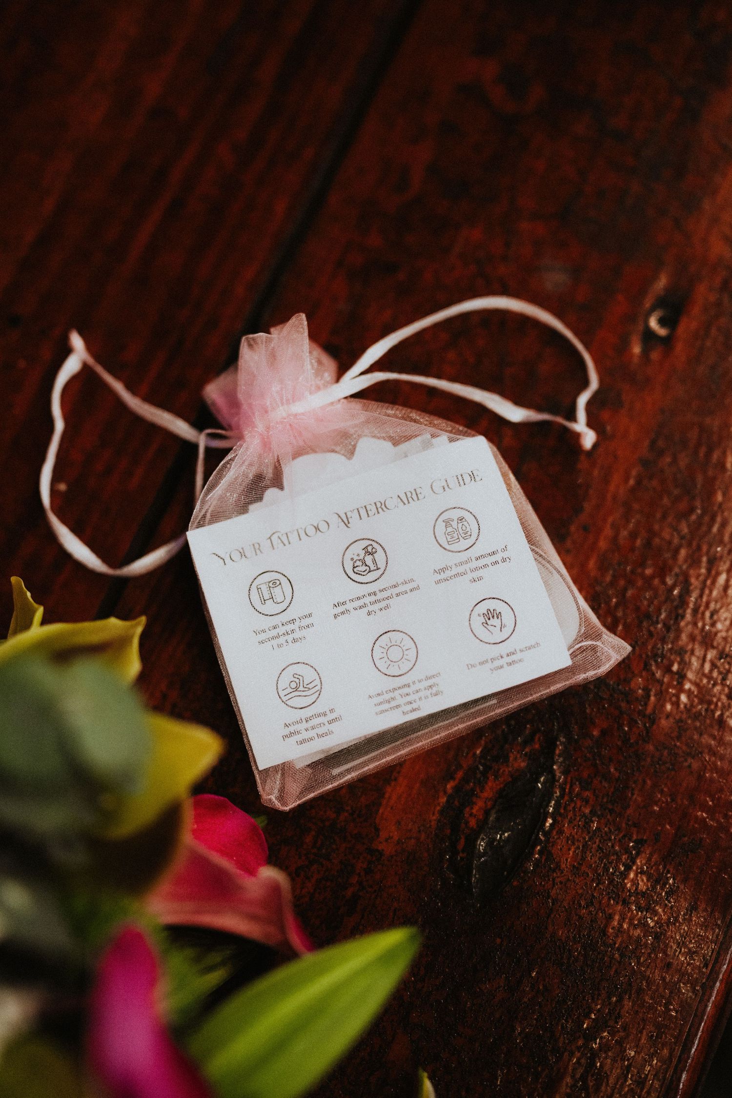 Pink organza bag with tattoo aftercare guide card on wooden surface, surrounded by colorful flowers.