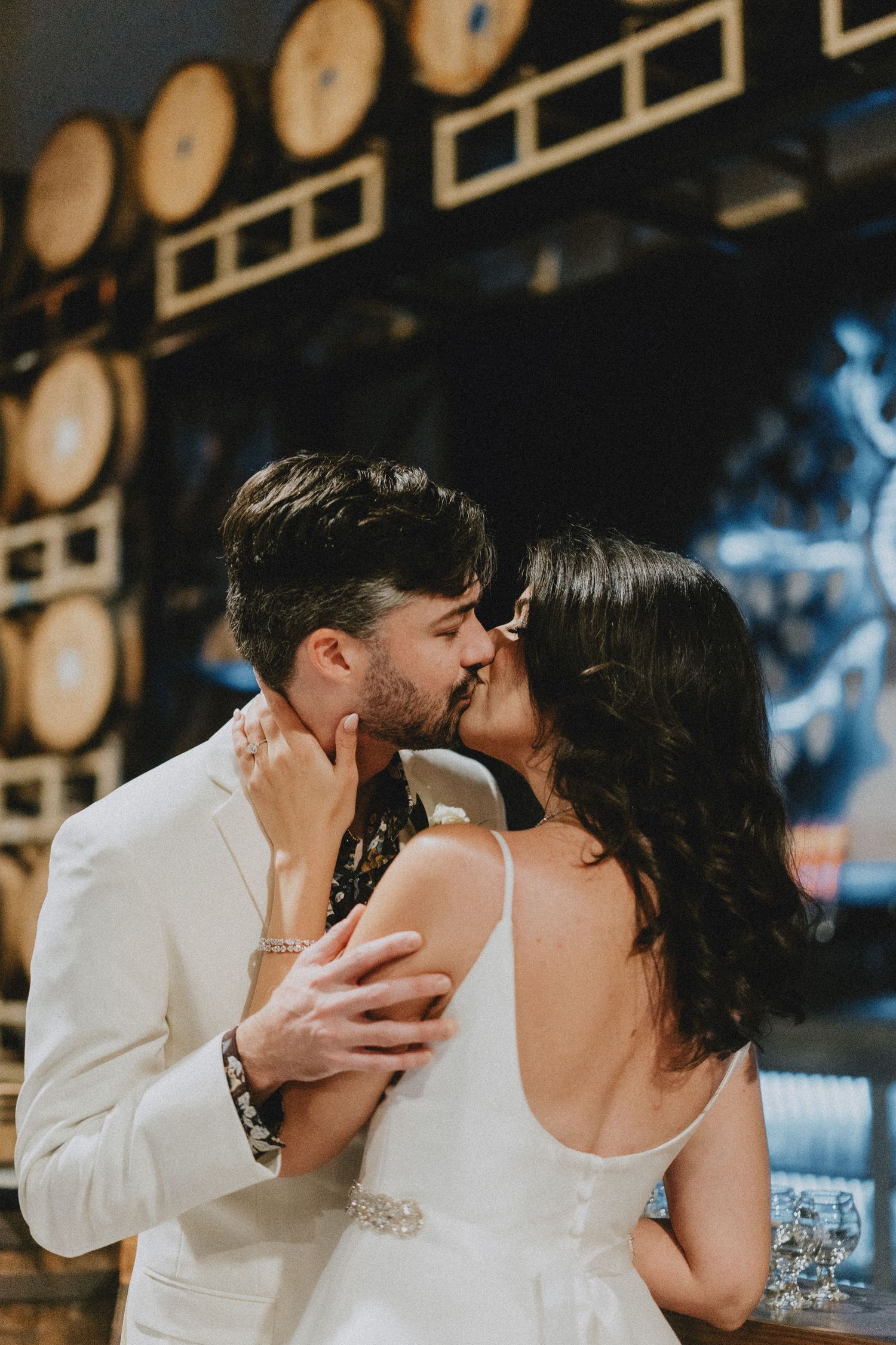 A couple shares a romantic kiss at their wedding, the groom in a white suit embracing the bride in a sleeveless gown.