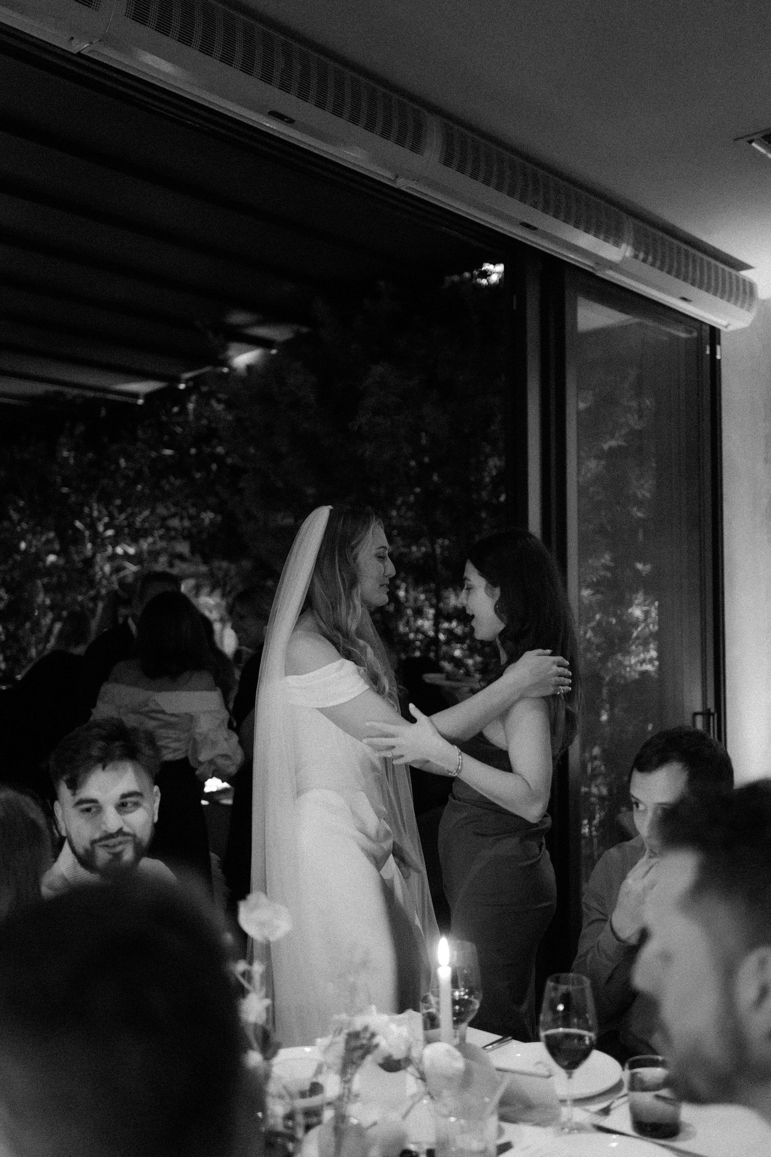 Black and white photo of bride in veil embracing female guest at candlelit wedding reception.