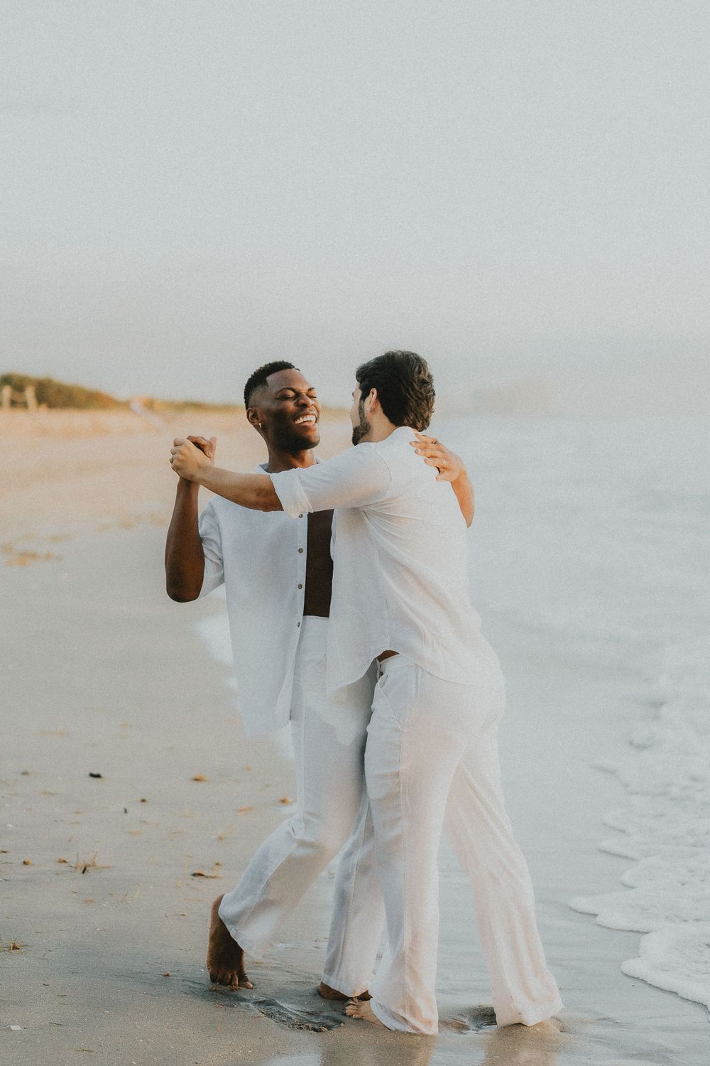 A couple in matching white outfits dancing and celebrating on a beach at sunset.