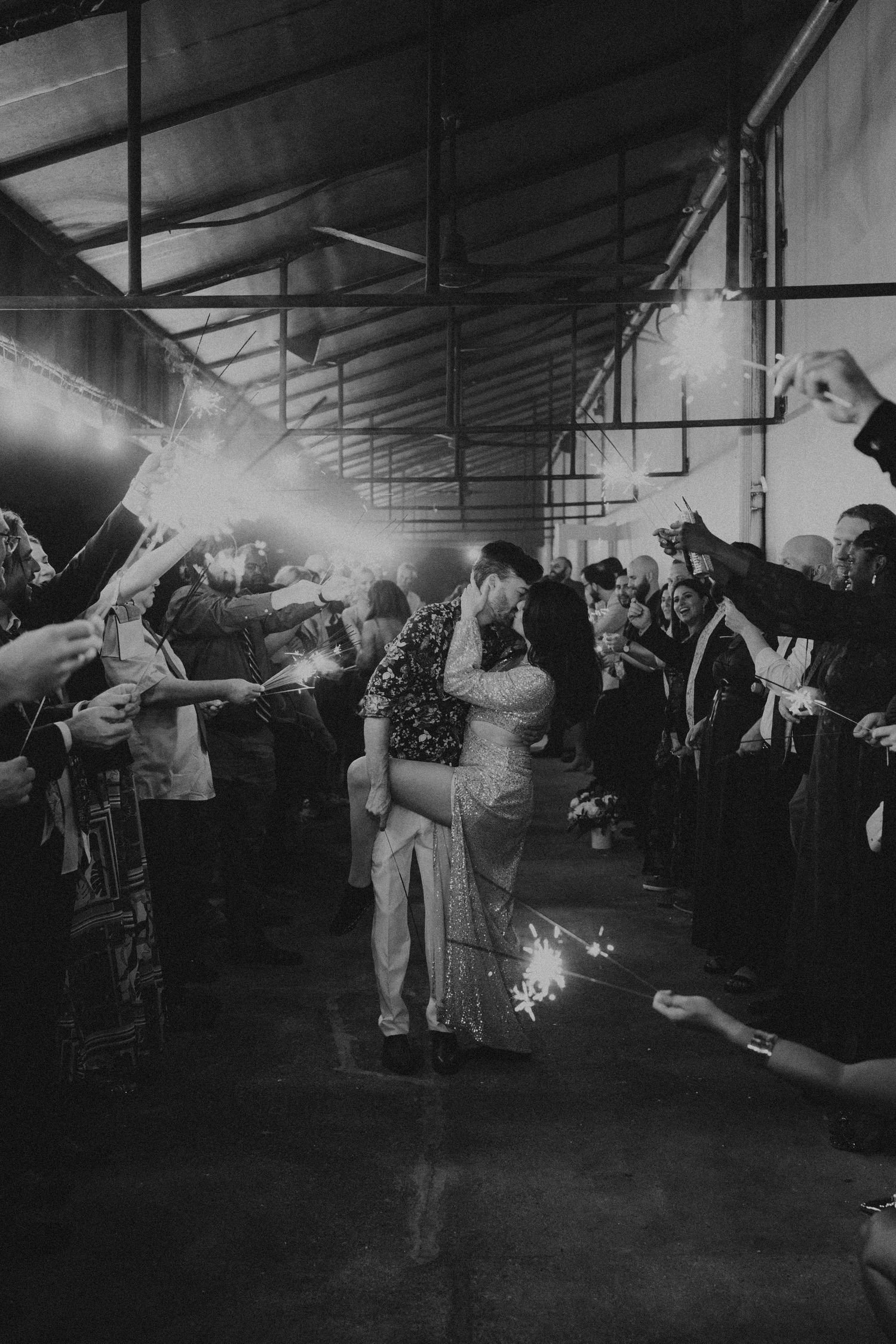 Black and white photo of newlyweds kissing during a sparkler send-off exit lined with cheering wedding guests.