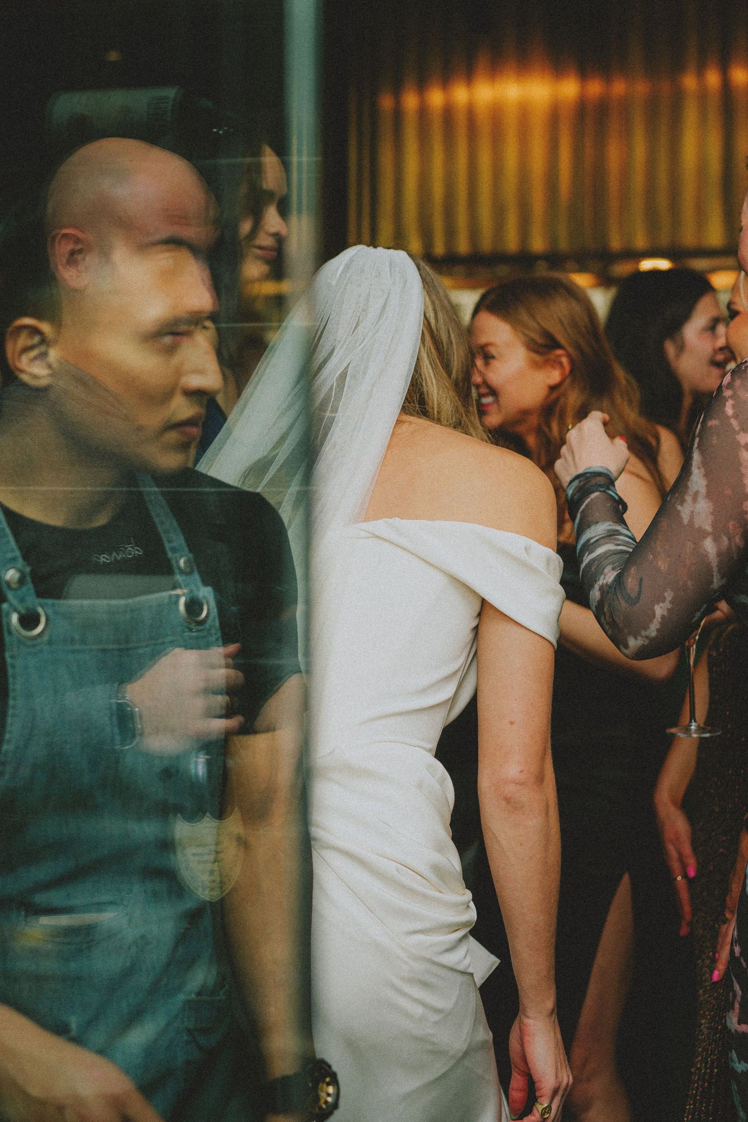 Bride in off-shoulder gown and veil embraces guest near reflective glass window at reception.