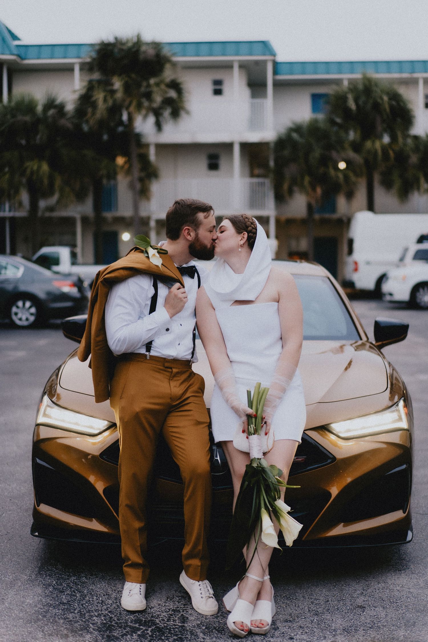 Newlyweds share a kiss leaning against a gold car after their wedding ceremony.