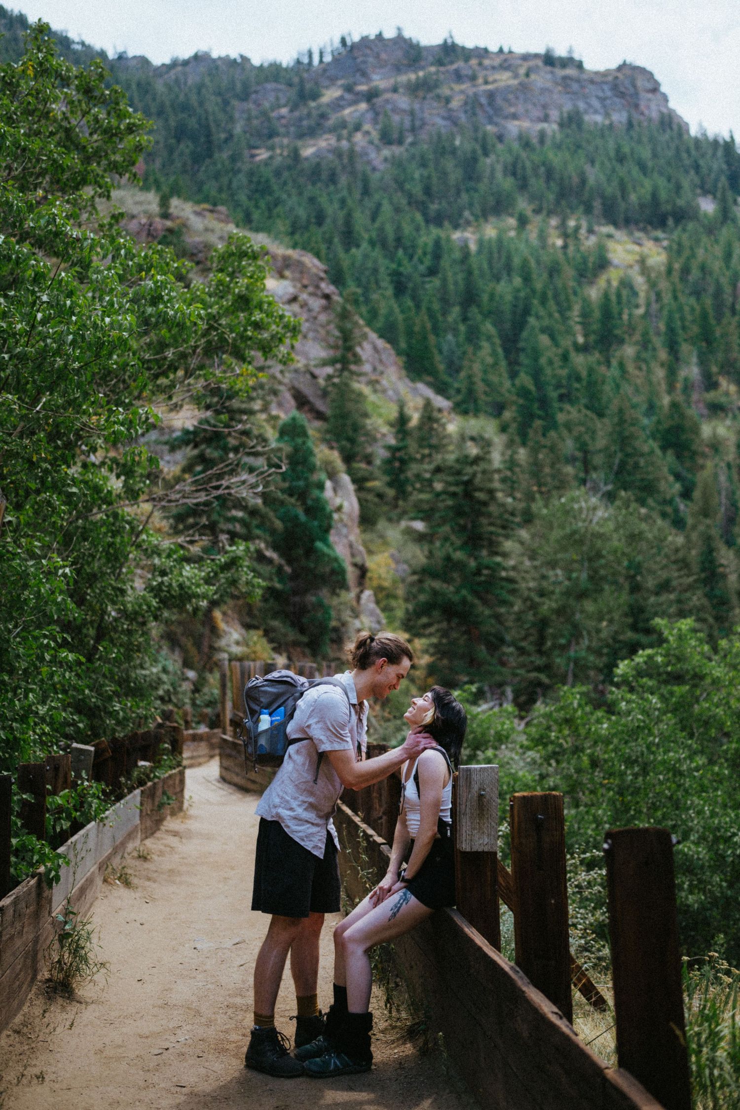 Couple sharing a romantic moment on a mountain trail bridge surrounded by lush pine trees and rocky cliffs.
