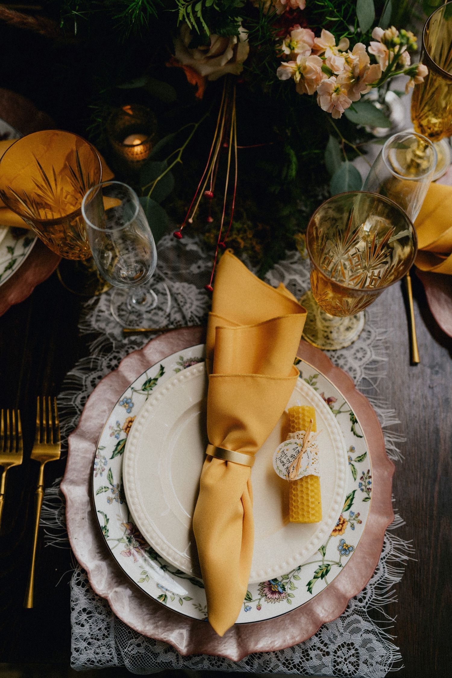 Elegant wedding table setting with mustard yellow napkin, floral china, gold cutlery, lace runner, and honeycomb candle accent.