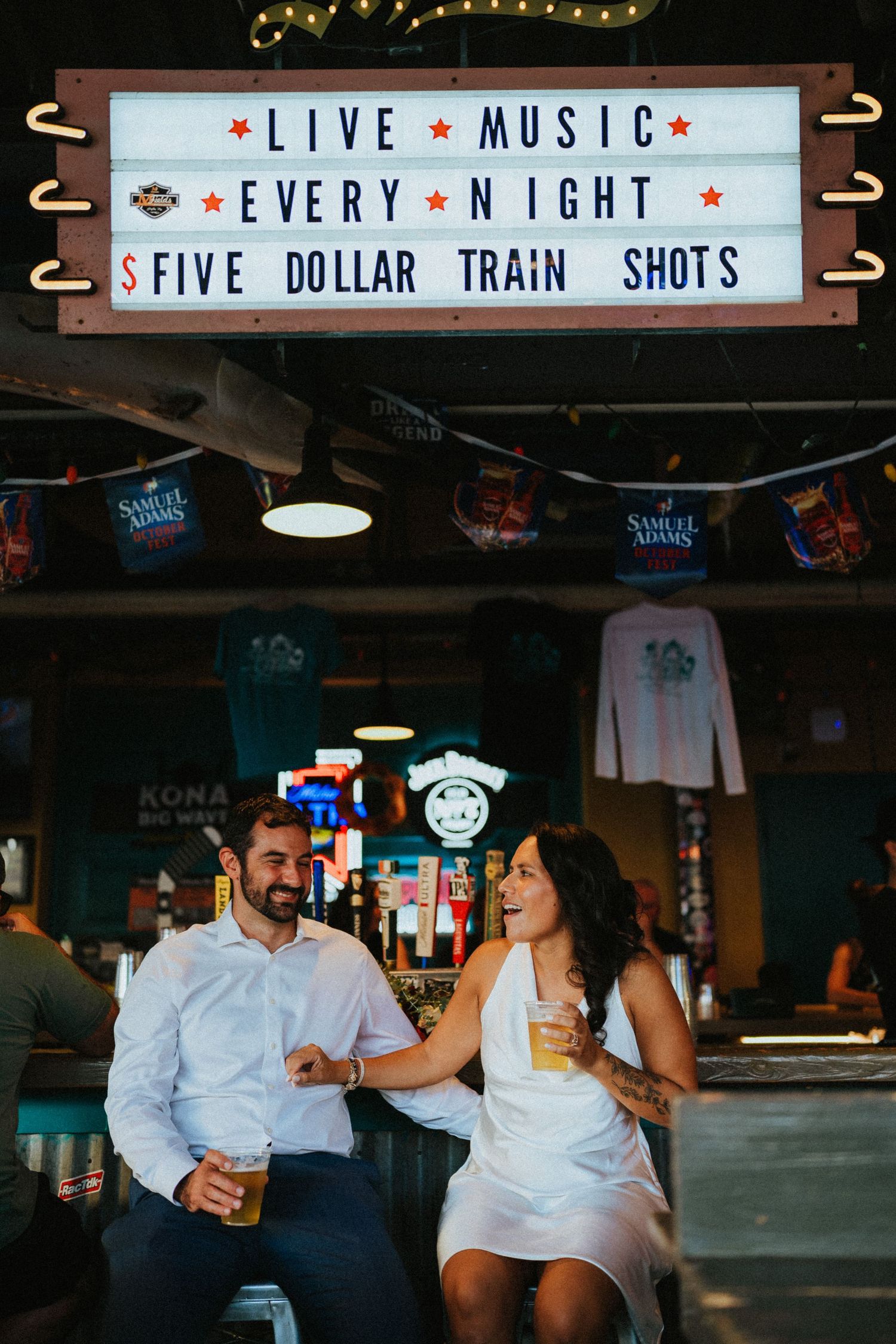 Couple laughing and holding beers at a bar under a live music and five dollar train shots sign.