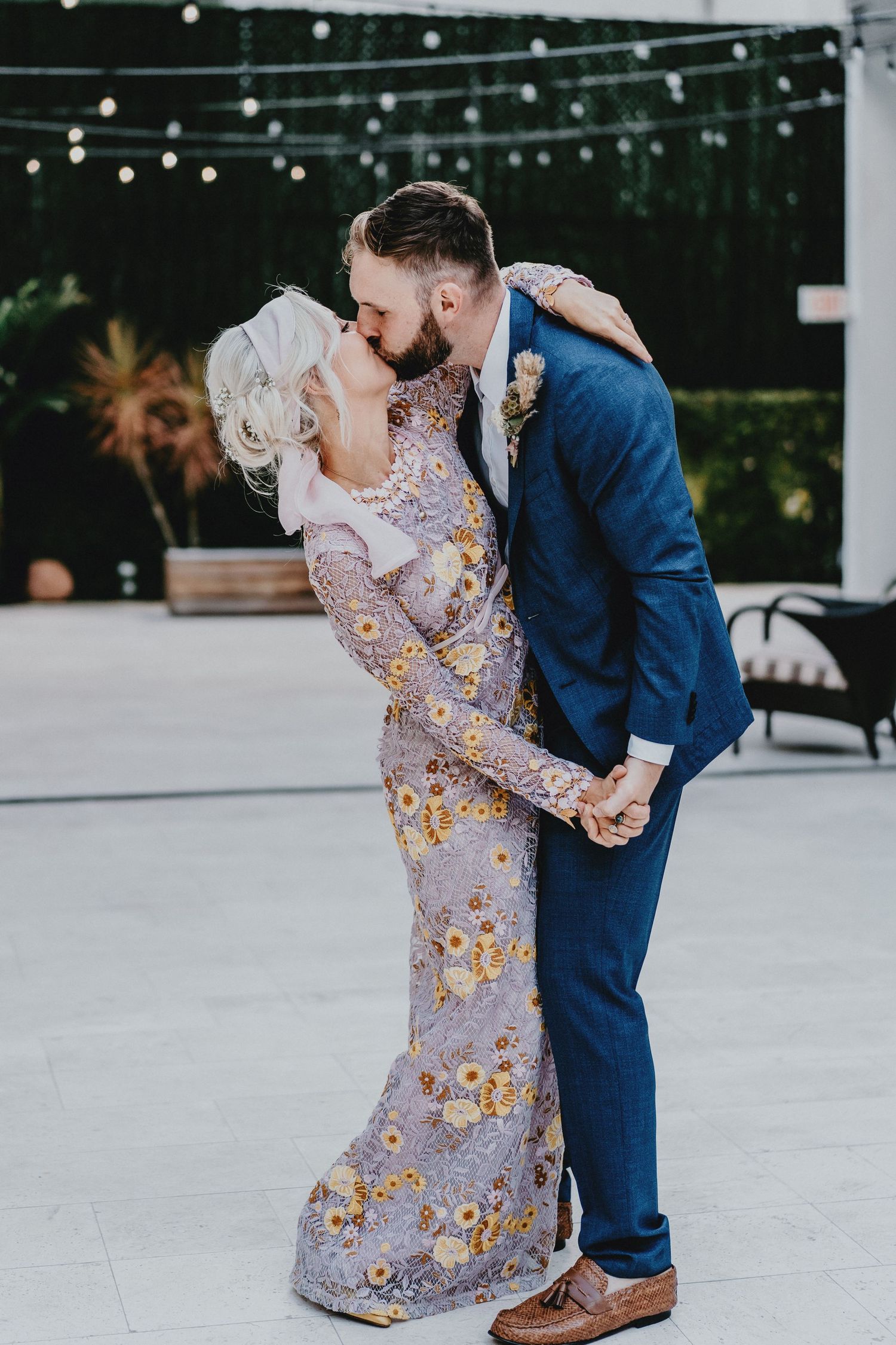 Bride in floral gown and groom in blue suit share a romantic dip kiss at outdoor wedding reception.