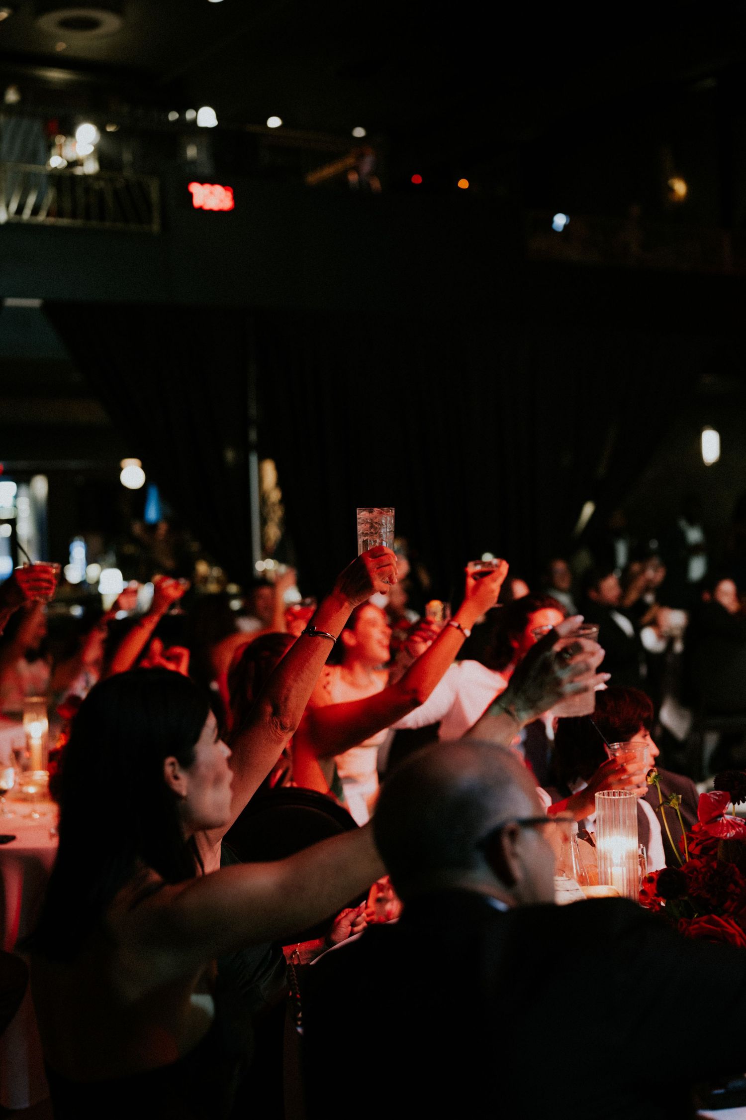 Wedding guests raising glasses in a toast during a dimly lit reception with red ambient lighting.
