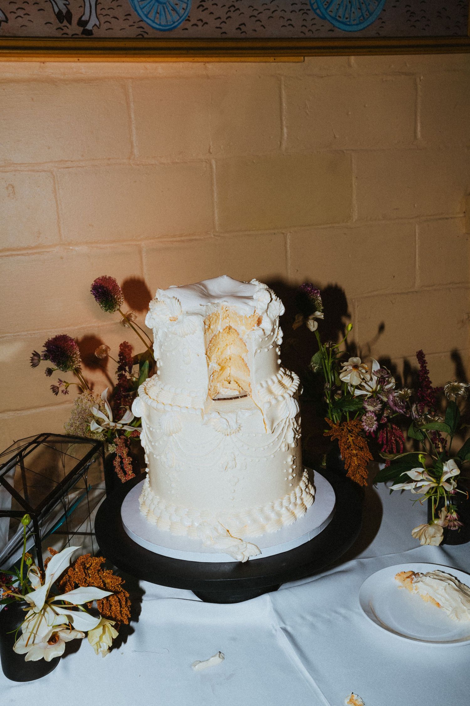 Partially sliced three-tier white textured wedding cake displayed on a black stand with wildflower arrangements.