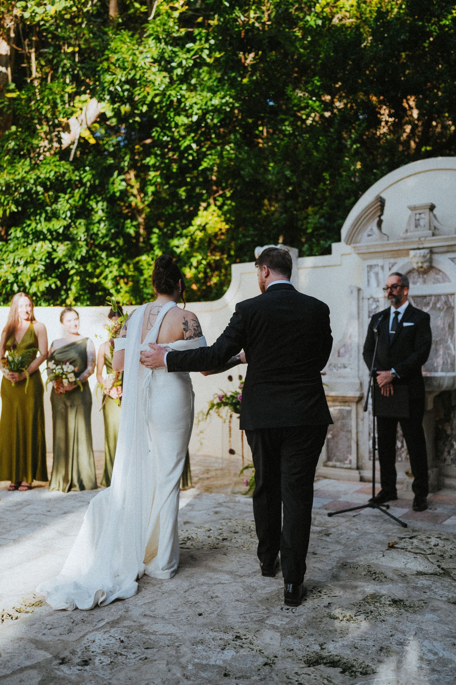 Bride and groom stand at outdoor wedding altar with bridesmaids in olive gowns and officiant beside ornate stone fountain.