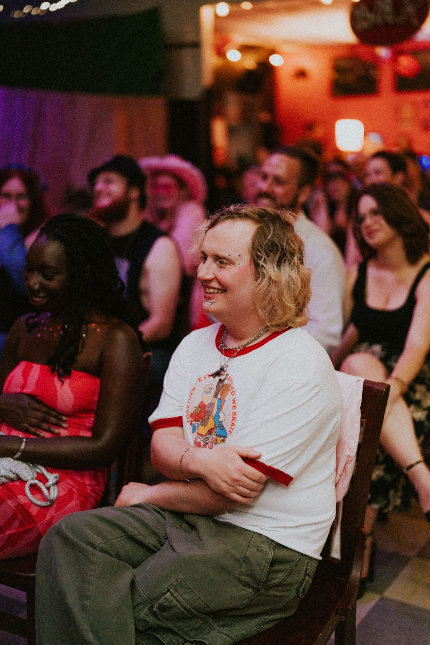 A smiling guest in a white ringer tee sits among a crowd of cheerful wedding attendees at a reception.
