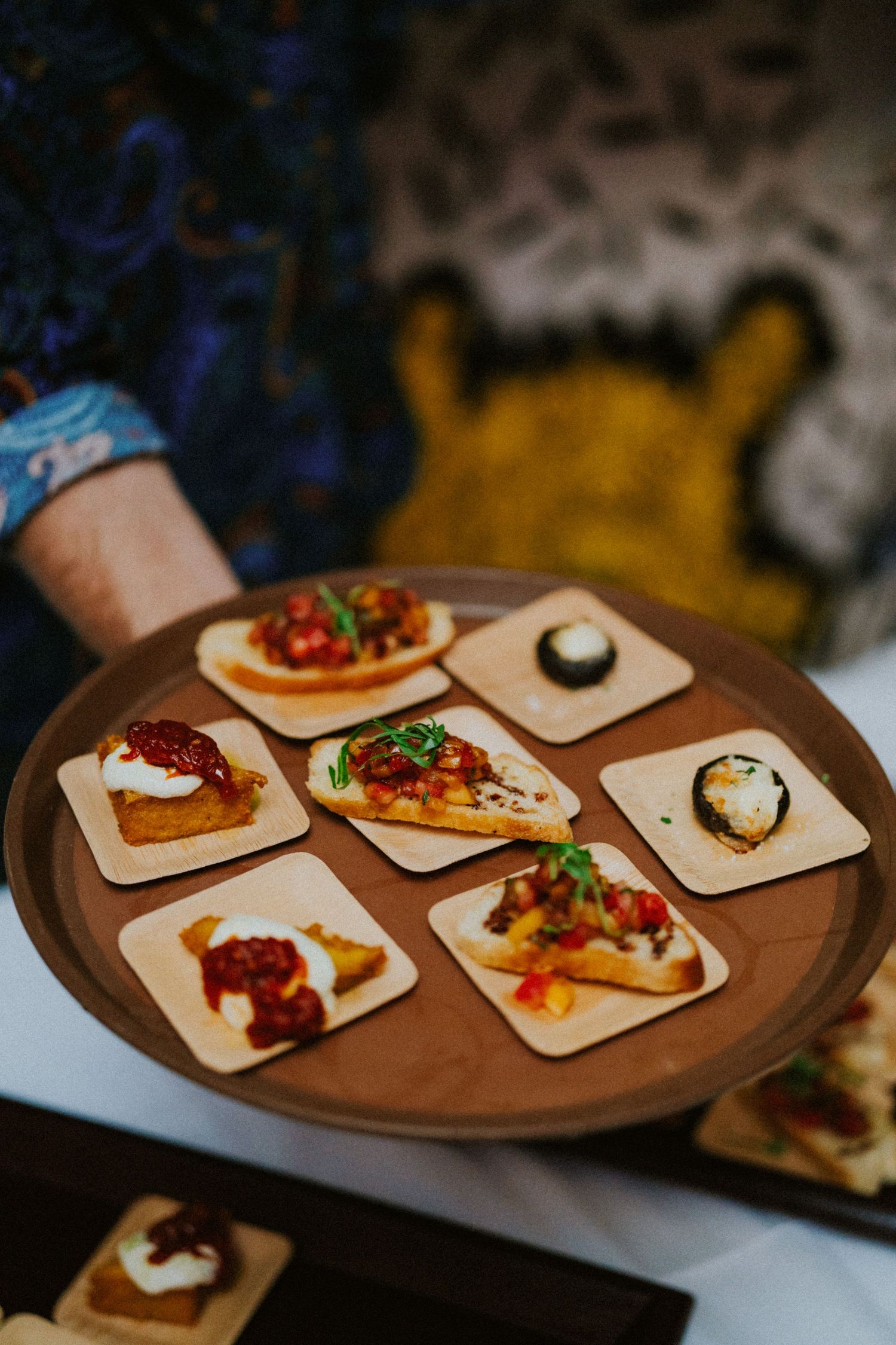 Server holding a wooden tray of elegant bruschetta appetizers topped with tomatoes and cream cheese at a catered event.