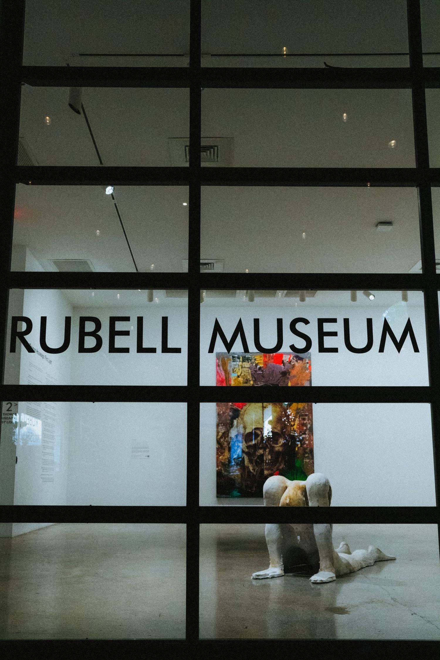 View through glass windows of Rubell Museum entrance with a sculptural figure crouching on the floor inside a gallery.