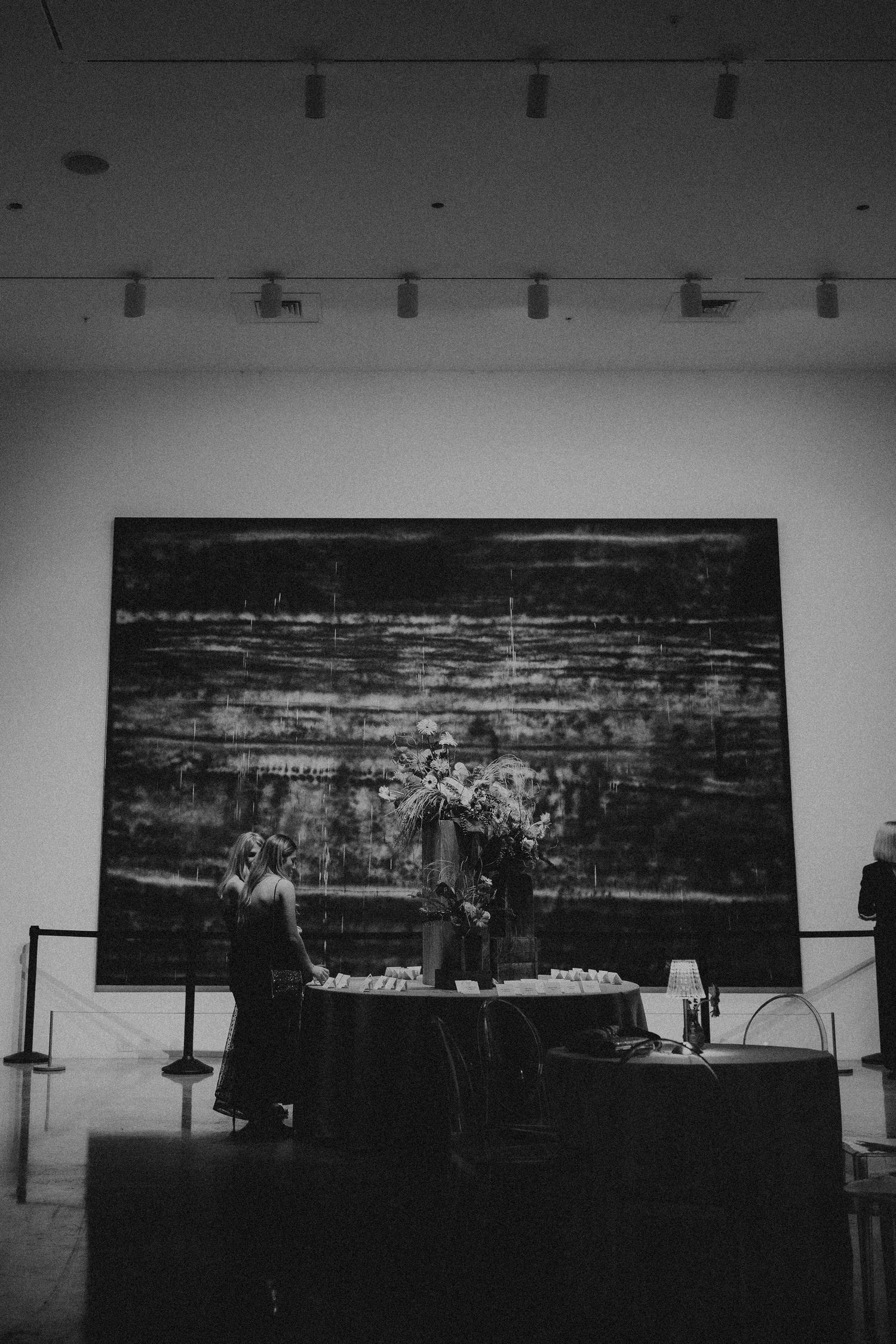 Black and white photo of a woman standing by a floral table display in front of a large abstract painting in a gallery.