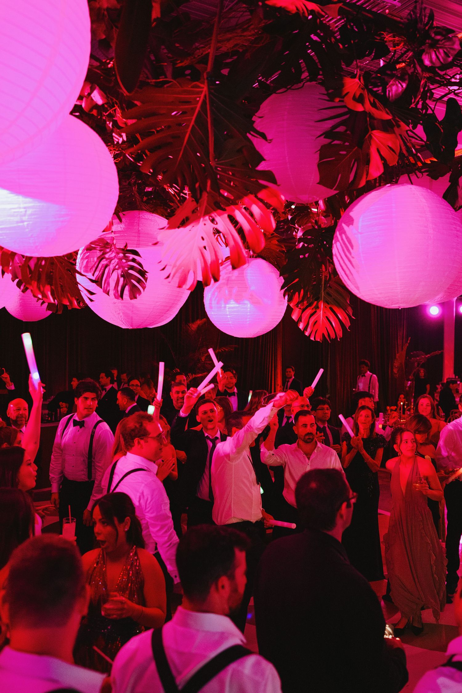 Elegant wedding reception with glowing pink lanterns, tropical greenery, and guests dancing with light-up wands.