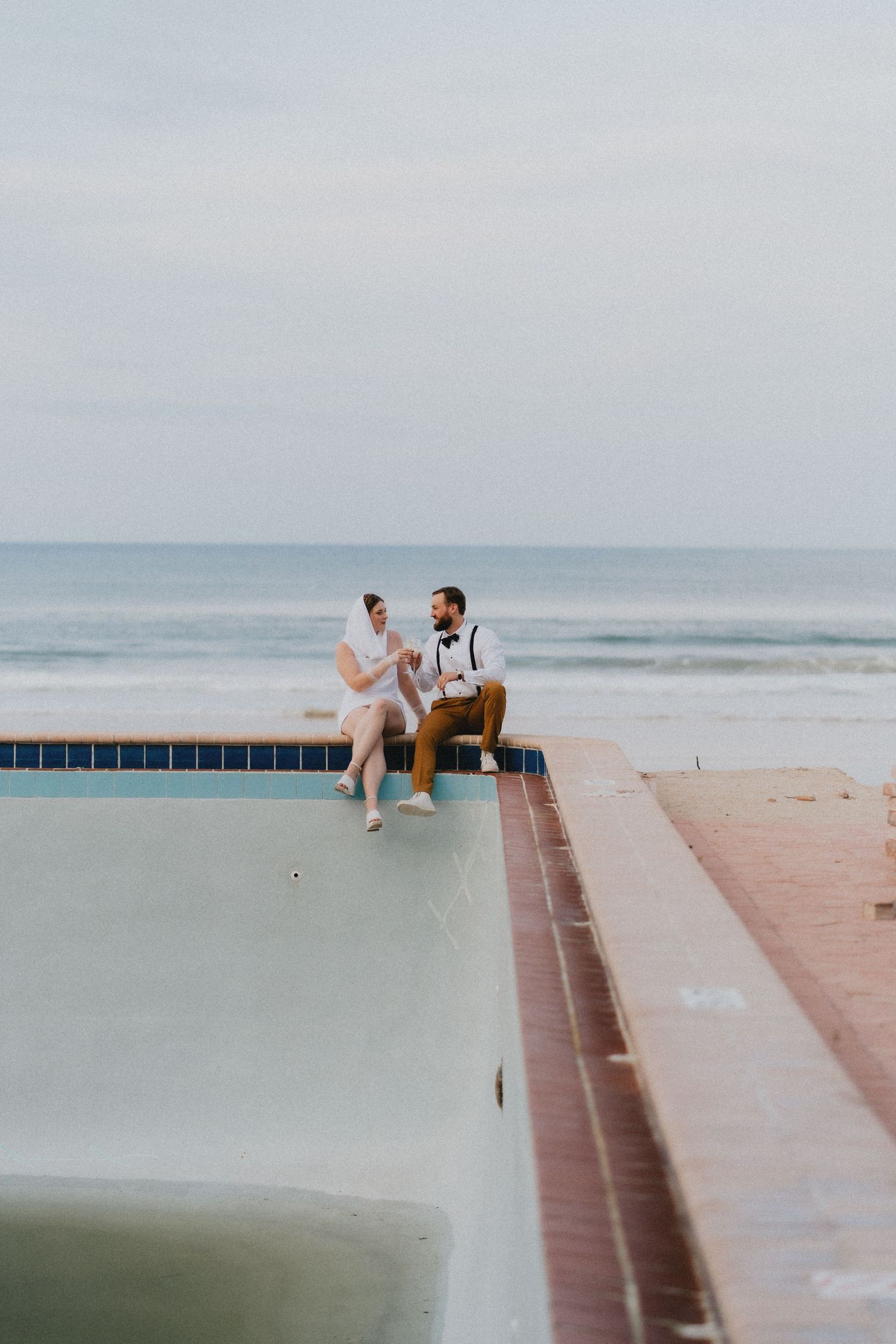 Bride and groom sitting on edge of a pool overlooking the ocean on their wedding day.
