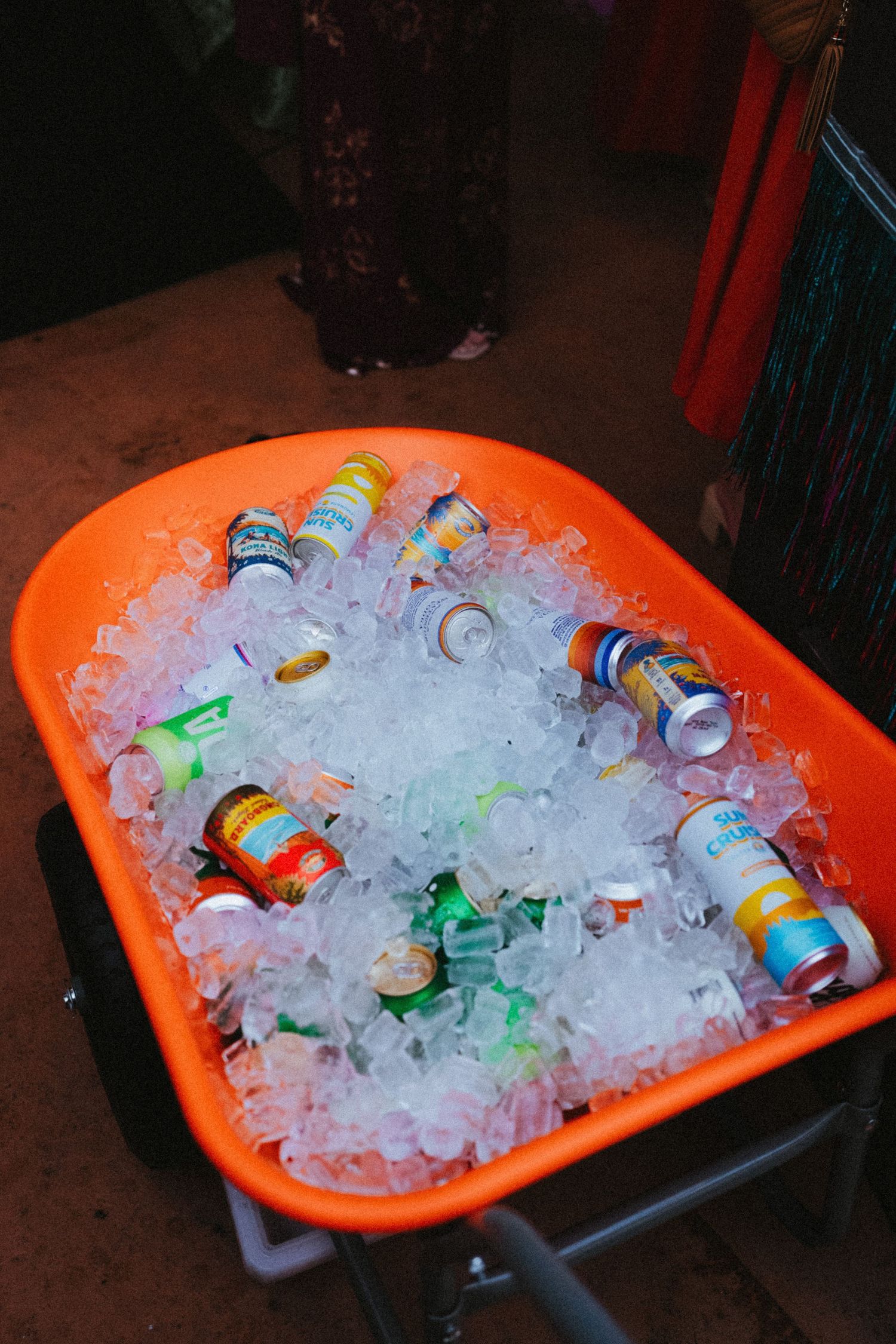 Orange wheelbarrow filled with ice and assorted canned beverages serves as a rustic drink cooler.